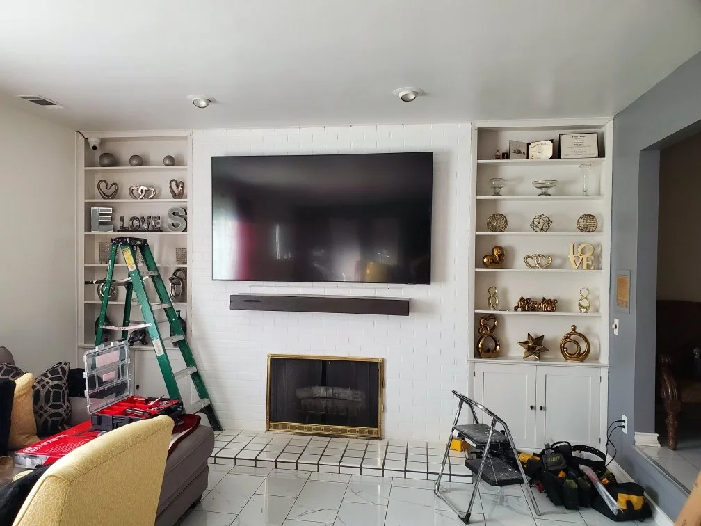 Home theater and sound system installed above a fireplace, between two in-wall bookshelves. A tall ladder and step ladder with a couple tool boxes are arranged in the living room in front of the home theater.