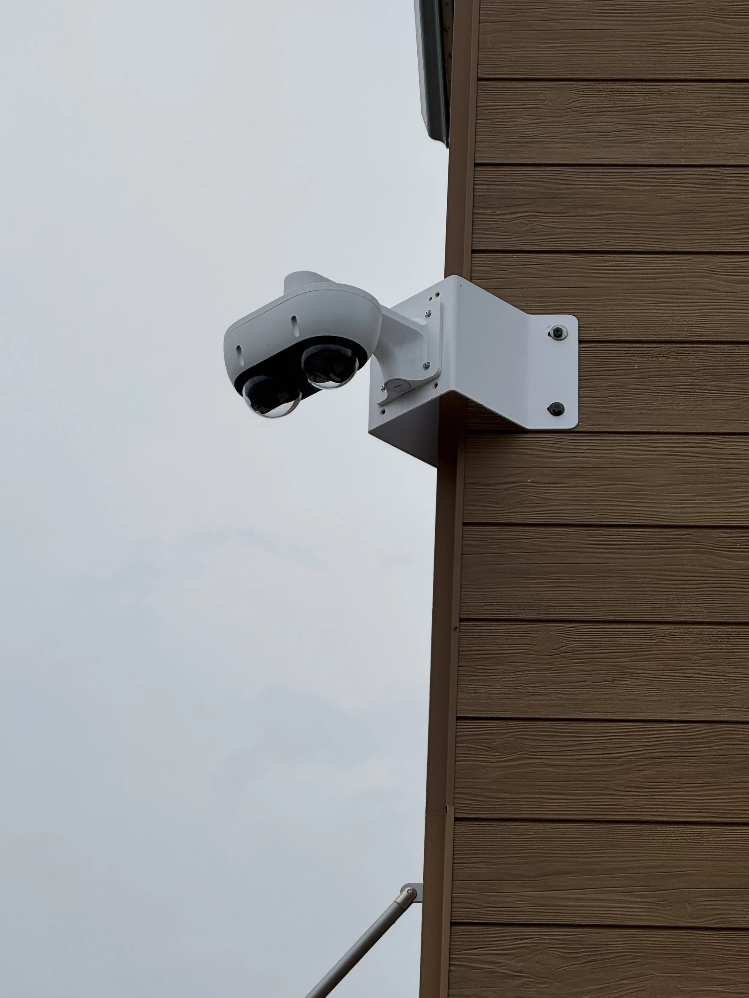 A white security camera attached to the corner of a brown wooden building with an overcast sky in the background.