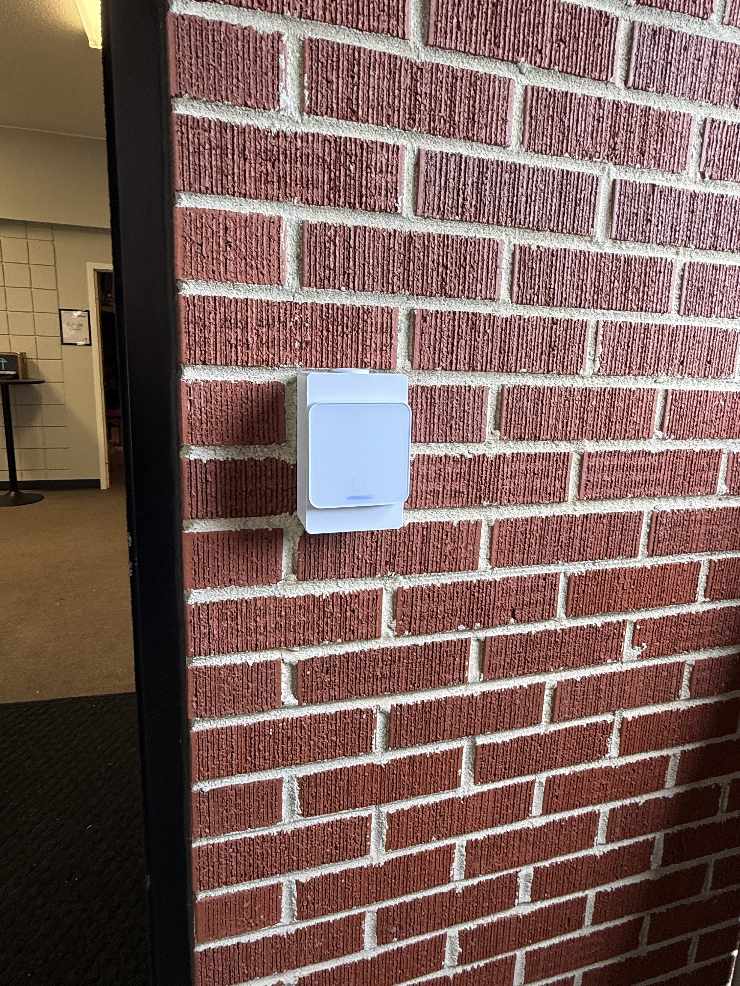 A brick wall with a white device attached to it, next to a black door frame. The interior of a building is visible in the background.