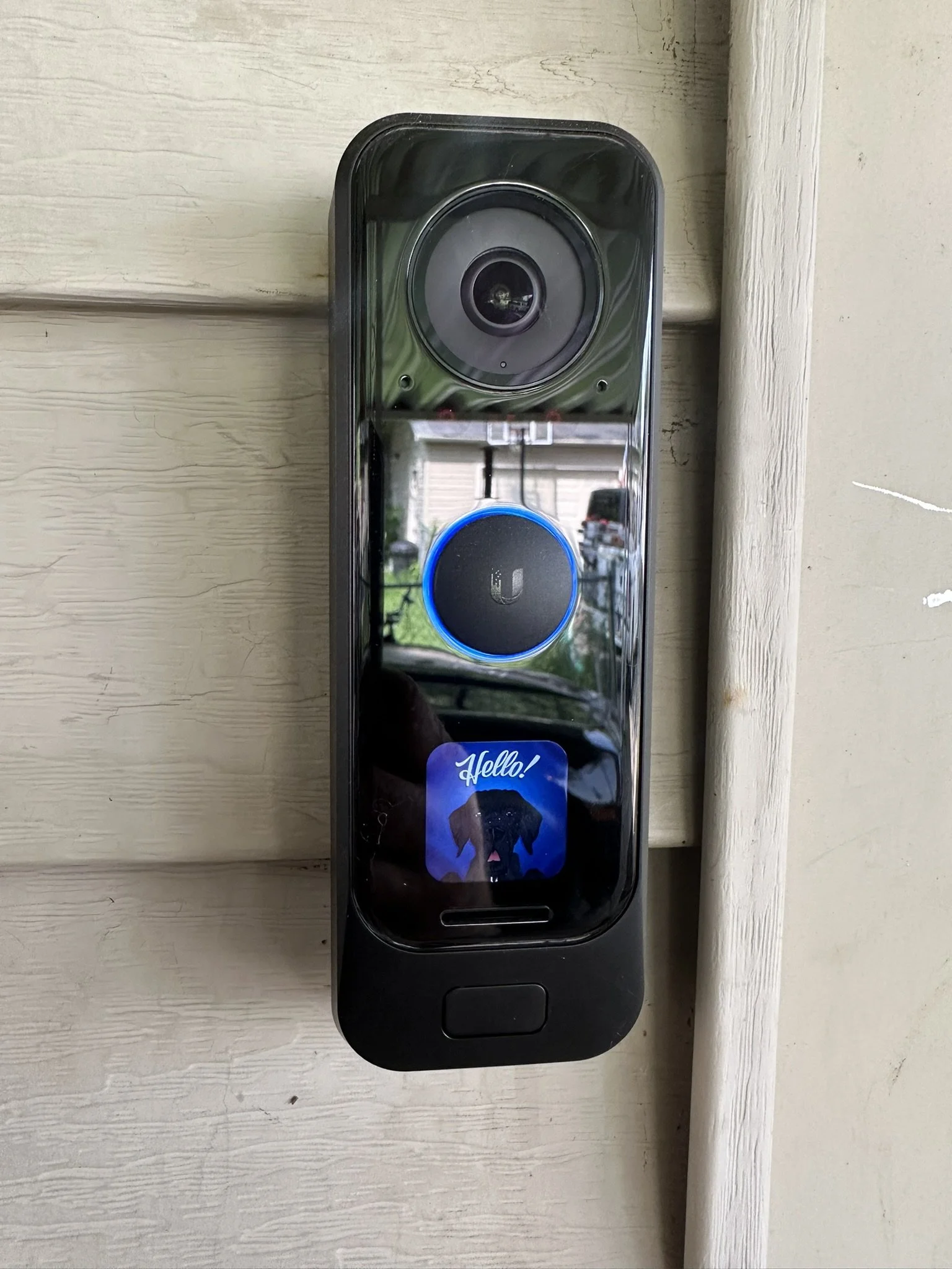 Doorbell camera with a blue circular button, a small sticker with a black dog and the word "Hello!", and a camera lens at the top, mounted on a beige wooden wall.