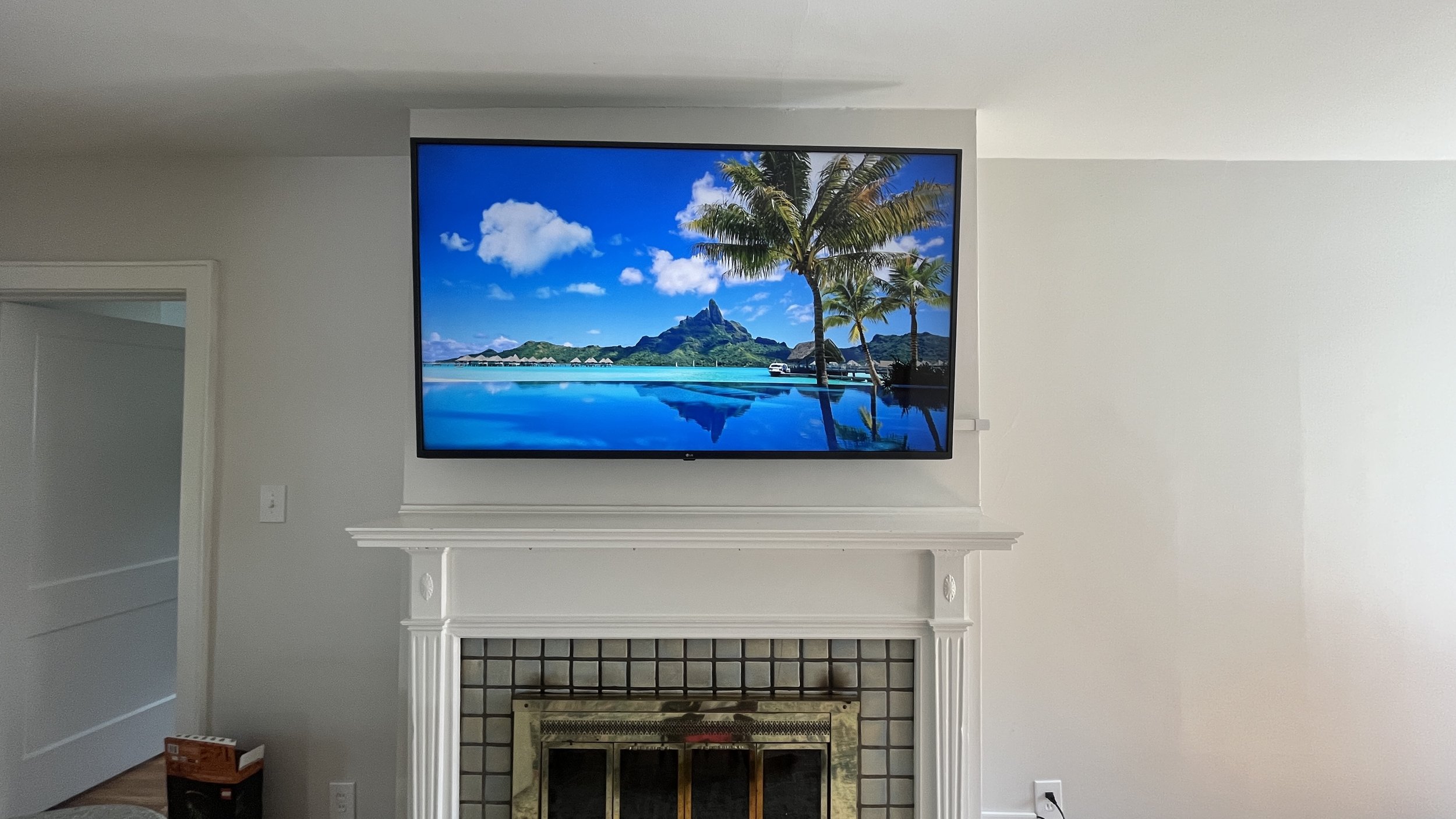 Living room with a white fireplace mantel and a flat-screen TV displaying a tropical beach scene with palm trees, mountains, and blue water.