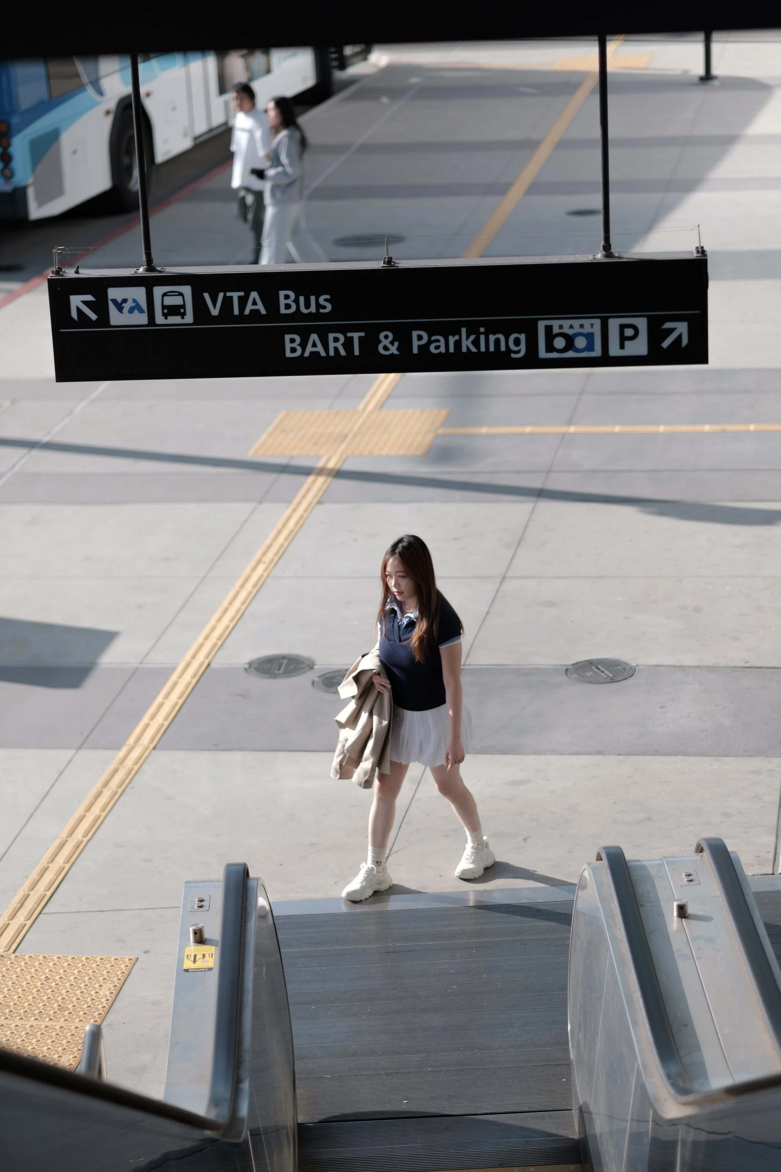 A young woman in a navy blue top and white skirt walking with a tan jacket in her hand at an airport or bus terminal, near escalators, with signs pointing to VTA bus, BART, parking, and bus services.