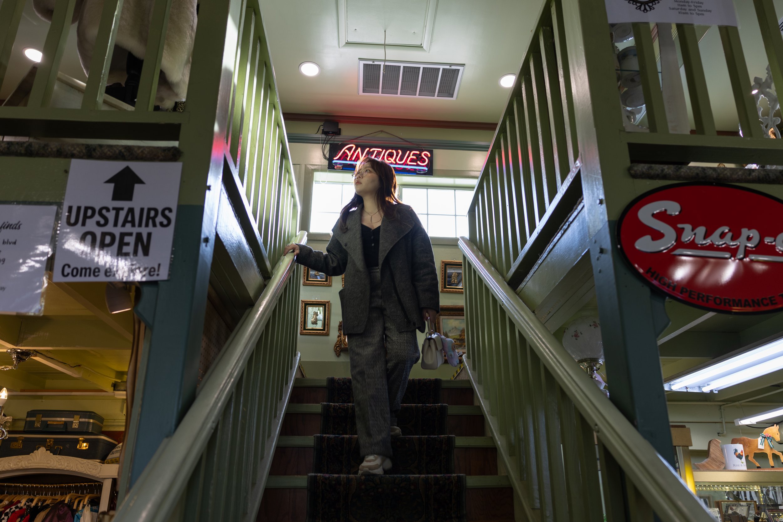 A woman in a gray coat and checked pants walking downstairs inside a thrift store. There is a neon sign reading 'ANTIQUES' behind her and signs indicating the upstairs is open. The store has framed artwork on the walls and various items on display.