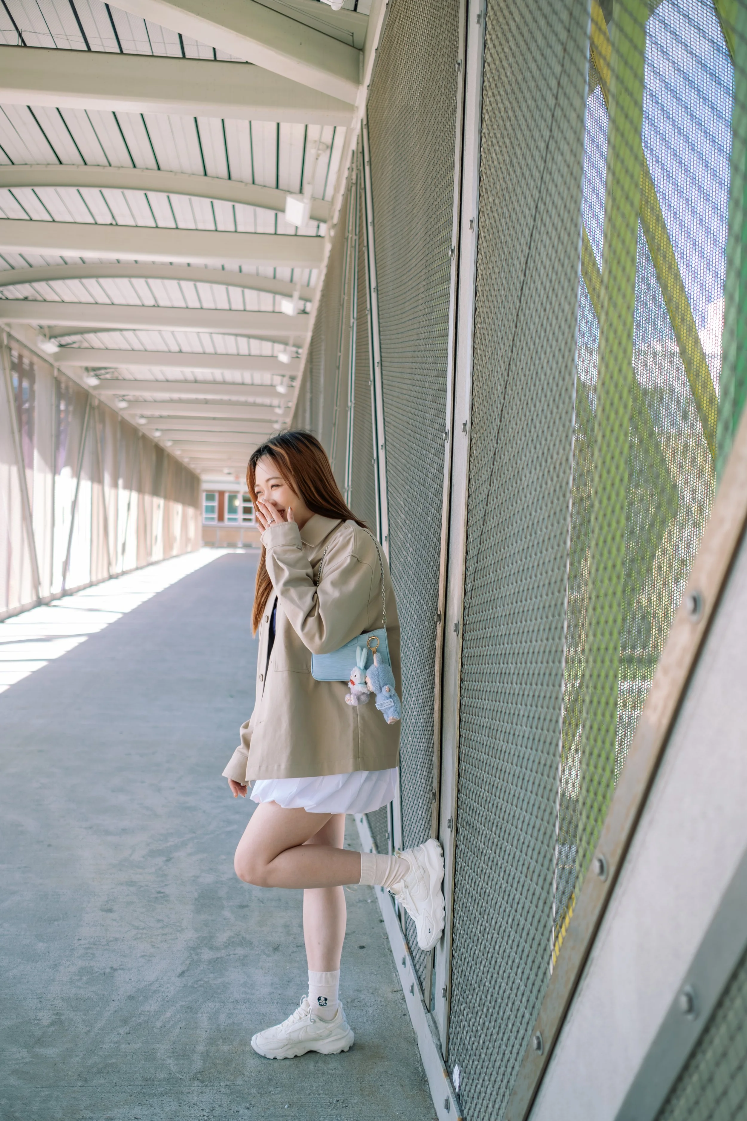 A young woman laughing and holding her hand to her face, standing beside a metal fence on a covered outdoor walkway, wearing a beige trench coat, white skirt, white sneakers, and a small blue handbag with plush charms.