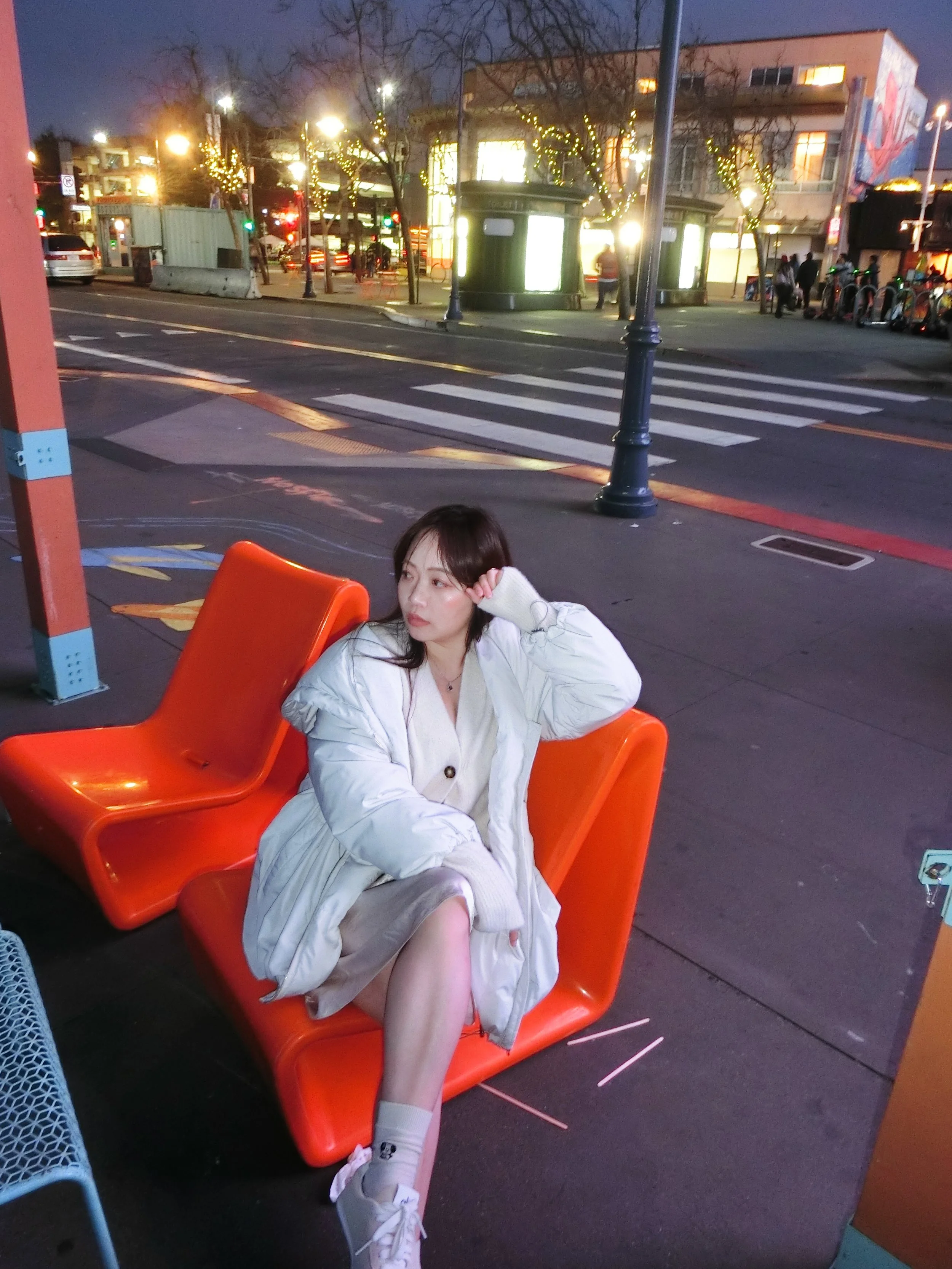 A young woman with dark hair sitting on an orange outdoor seat at a city bus stop in the evening. She is wearing a light-colored coat, a beige dress, white socks, and sneakers. She appears to be lost in thought, resting her head on her hand. The back