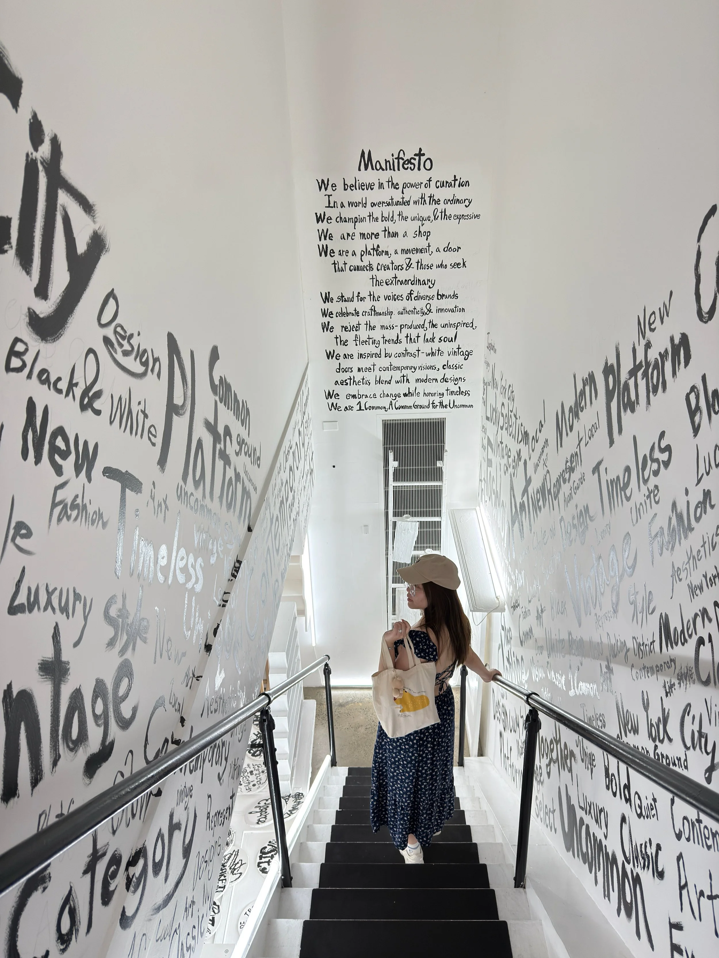 A woman with a beige cap, sunglasses, and a dark blue dress with white pattern, carrying a beige tote bag, walking down a staircase with black steps and white sides. The staircase walls are decorated with black and white handwritten words and phrases