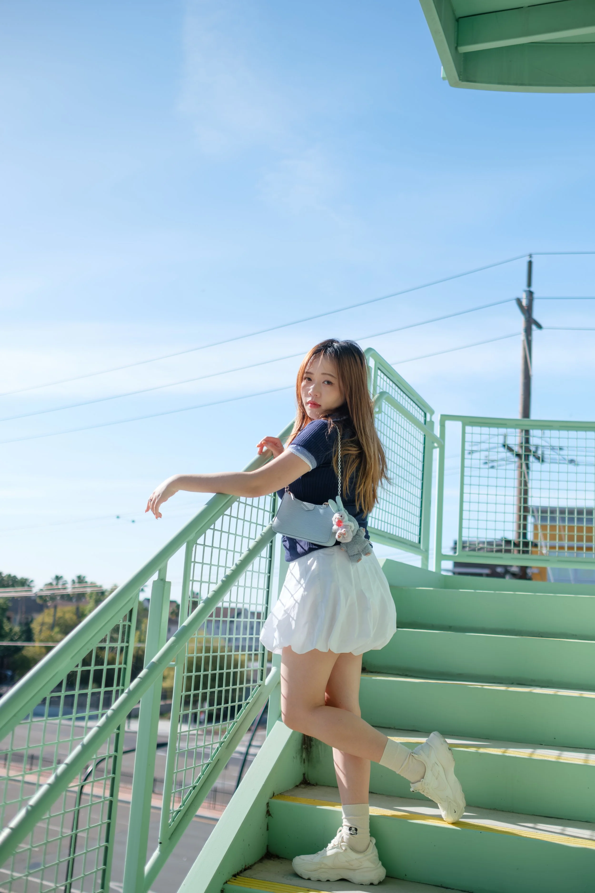 Young woman with long hair, wearing a navy blue shirt, white skirt, and white sneakers, stands on light green outdoor staircase against a blue sky with power lines.