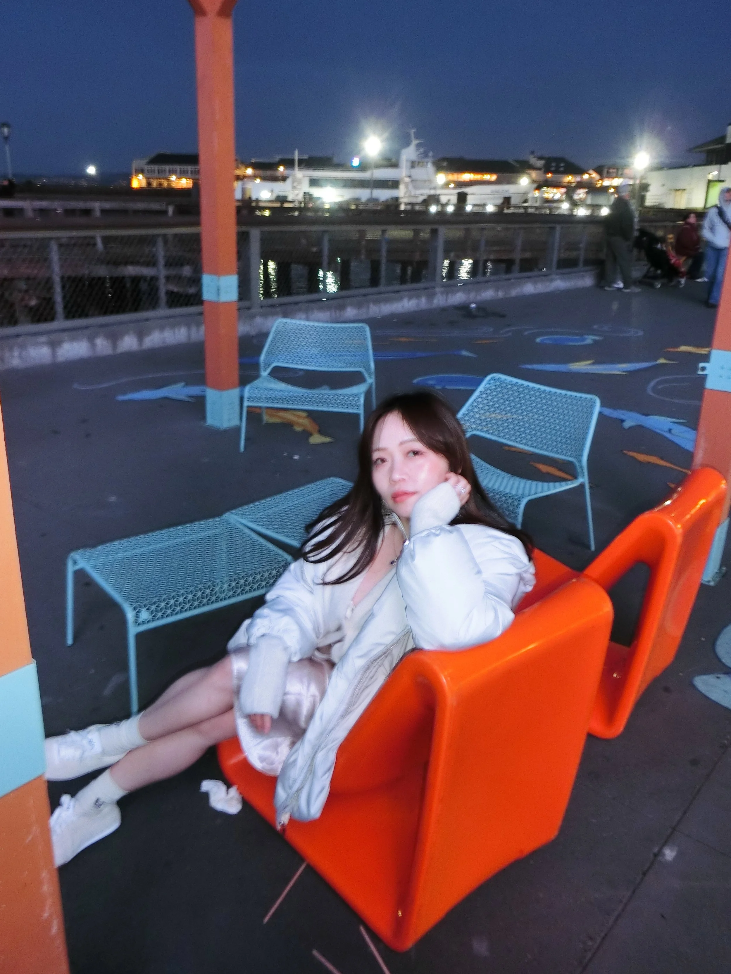 A young woman sitting on an orange lounge chair at an outdoor seating area near the water during dusk, with blue chairs around her and boats illuminated in the background.