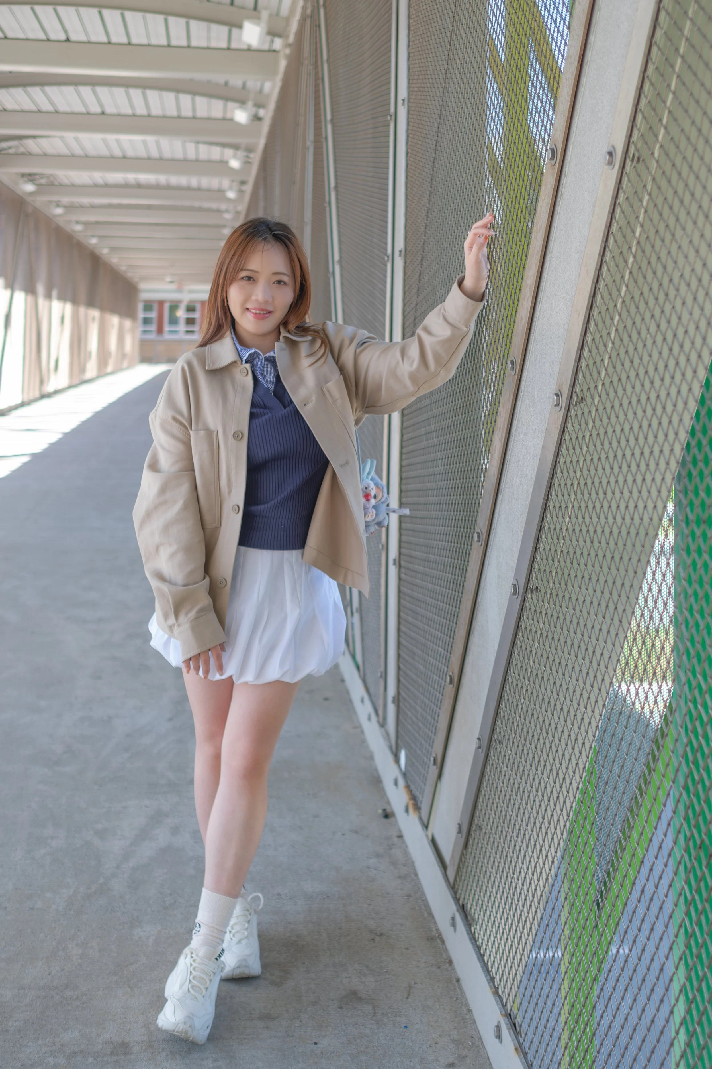 A young woman winking and smiling, dressed in a beige jacket, blue sweater, white pleated skirt, white socks, and sneakers, standing beside a metal fence under a shaded bridge or walkway.