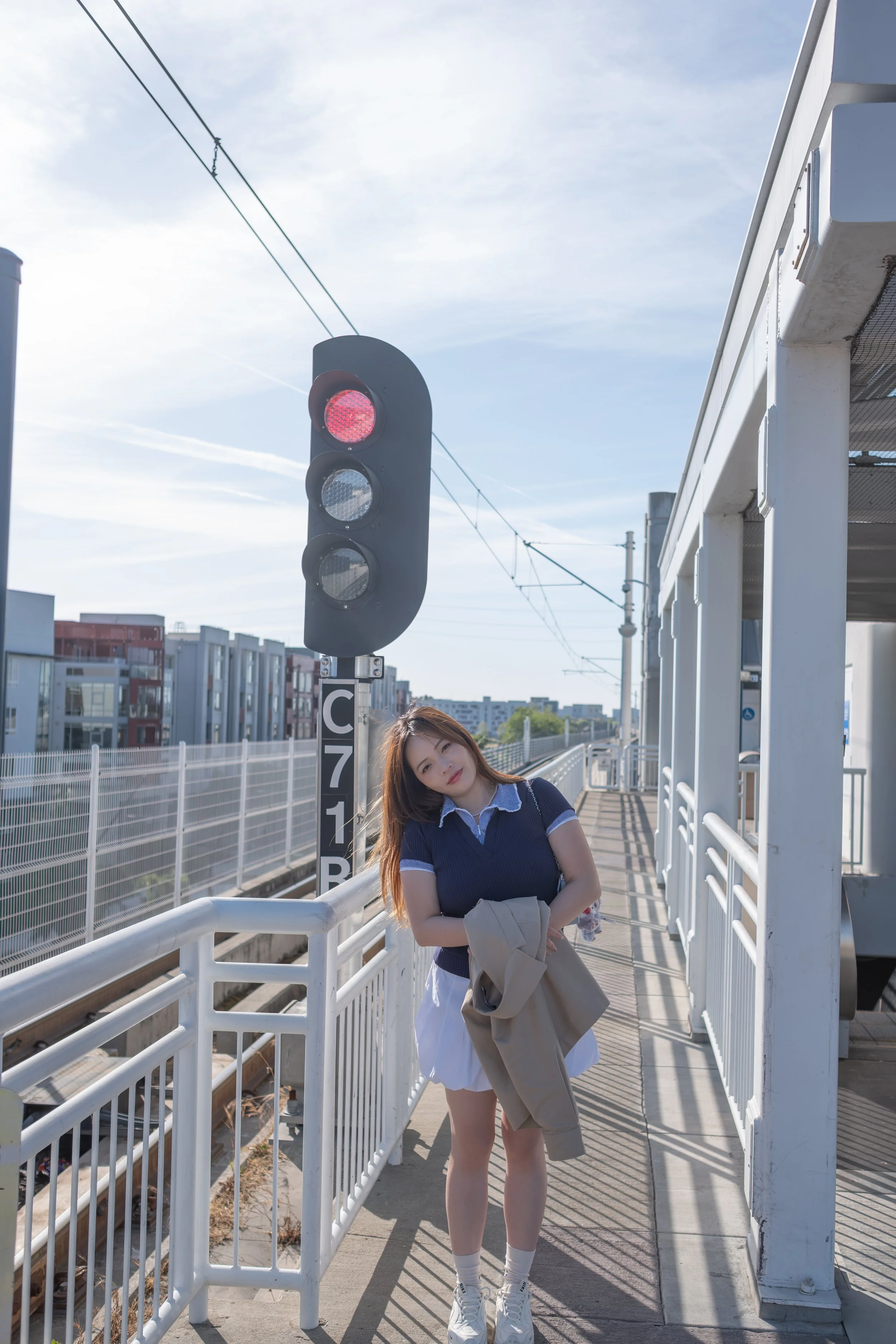 Young woman standing on train platform, holding a jacket, near a red warning light and train tracks, with modern buildings in the background.