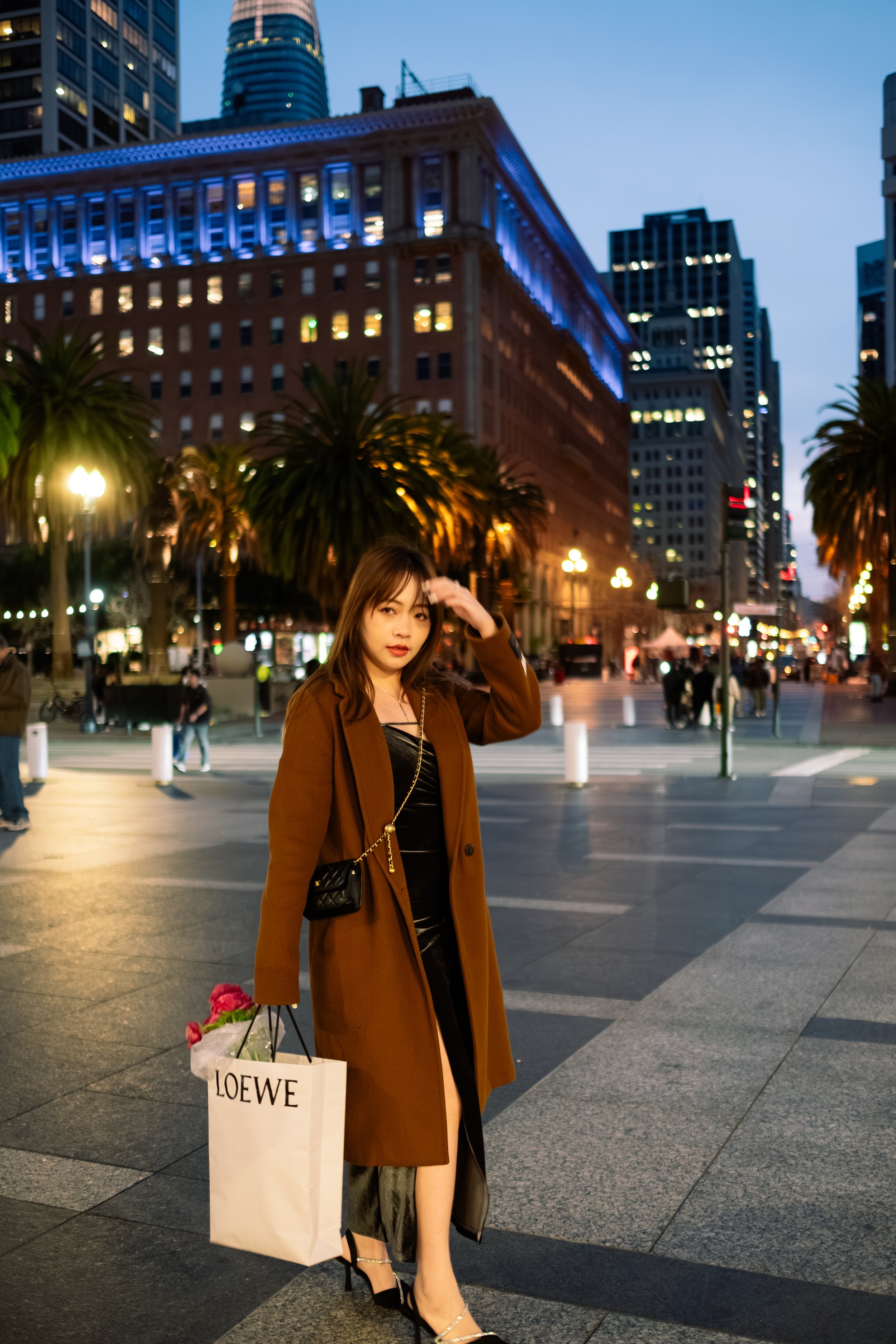 A young woman in a brown coat and black dress standing on a city street at dusk, holding a shopping bag from LOEWE with flowers inside, city skyscrapers with lights on behind her.
