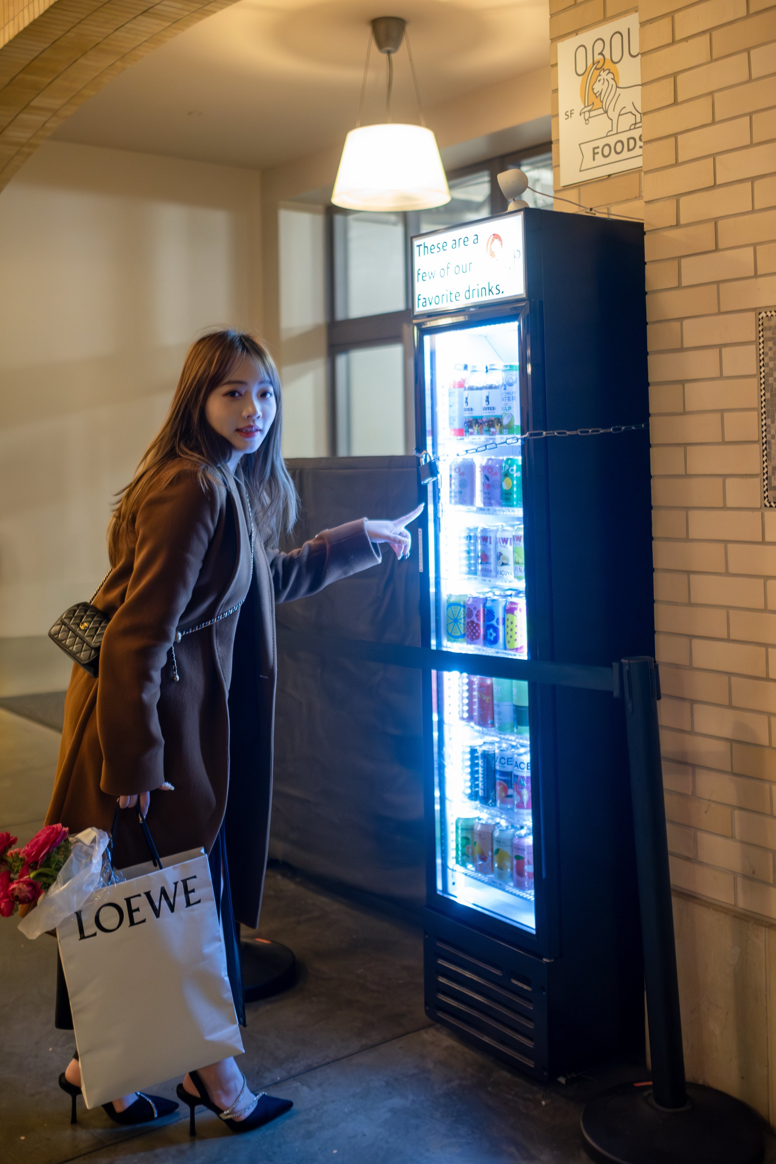 A young woman in a brown coat and black high heels is pointing at a vending machine that has a sign above it reading, 'These are a few of our favorite drinks.' She is holding a shopping bag with 'LOEWE' printed on it and a bouquet of pink flowers.