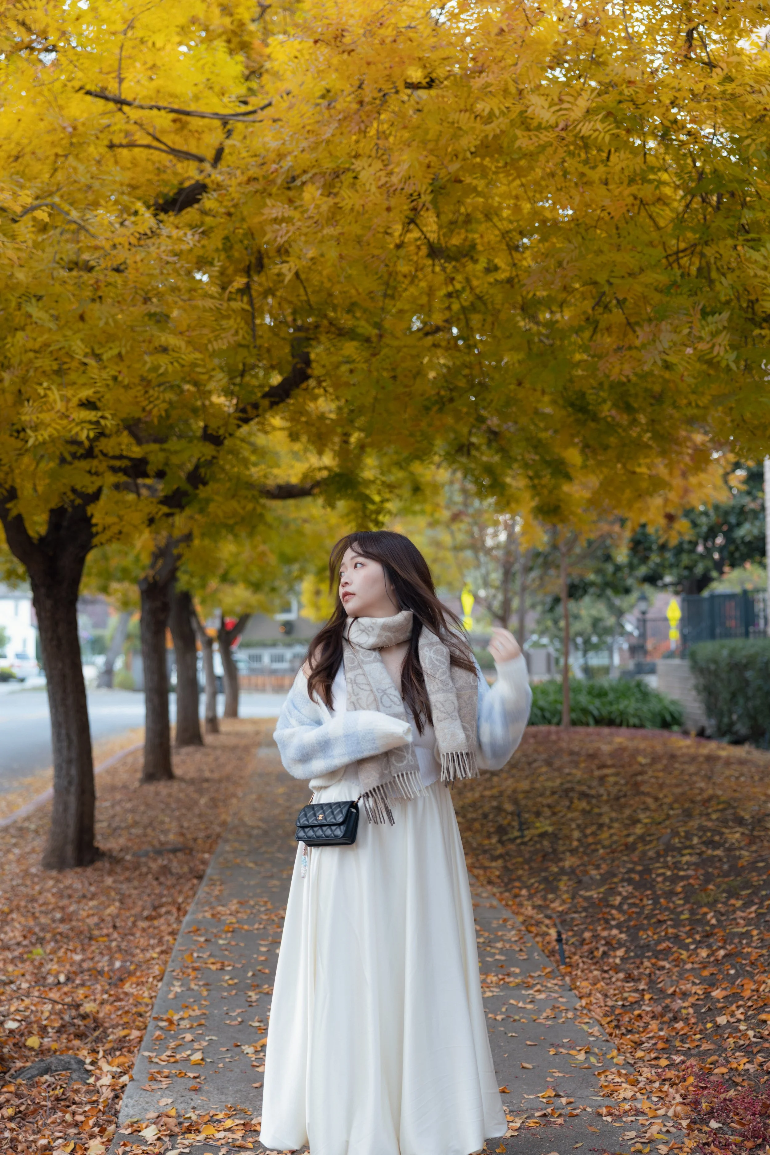 A woman in a long white dress and a cozy sweater with a patterned scarf standing on a fallen leaf-covered sidewalk under vibrant yellow autumn trees.