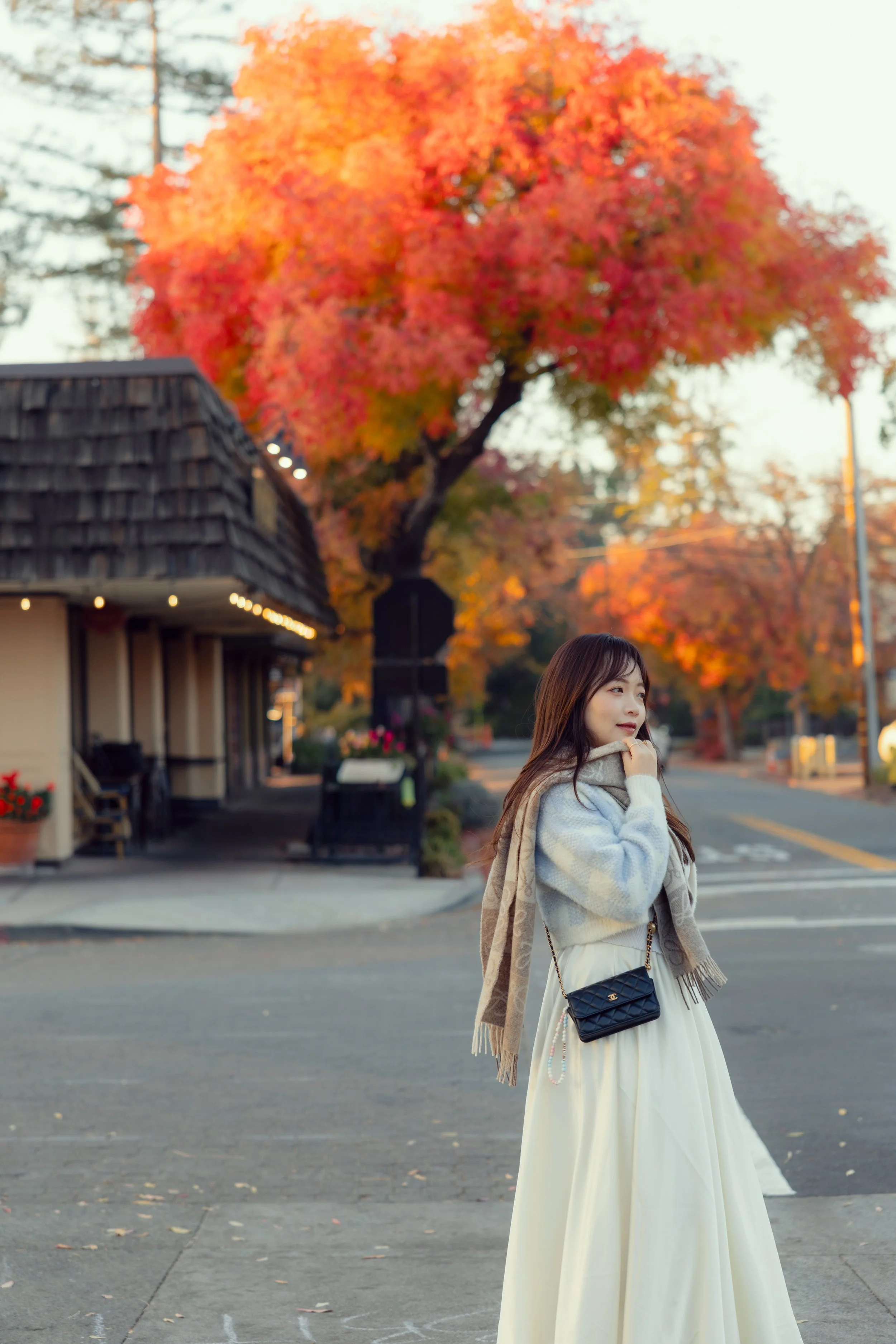 A woman in a white dress and beige scarf walking on a sidewalk in autumn with colorful fall leaves on trees in the background.