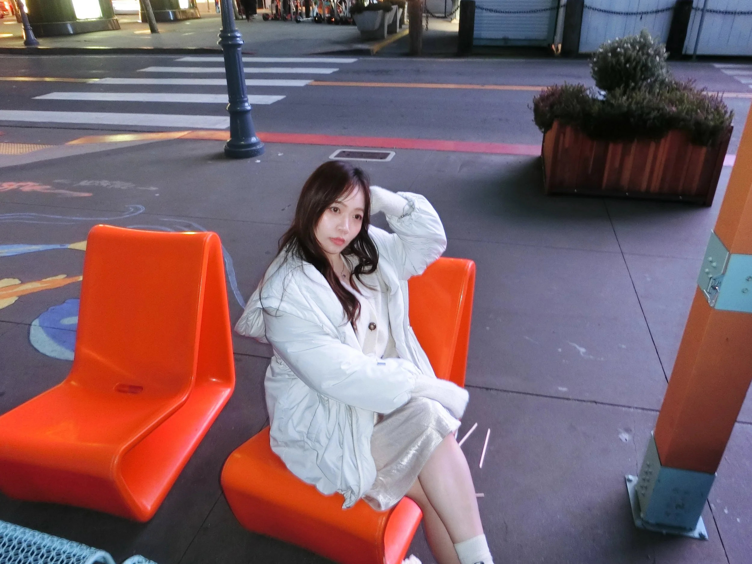A young woman with long hair sitting on an orange bench outside, dressed in a white jacket and light-colored dress, near crosswalk and street with trees and planters in the background.