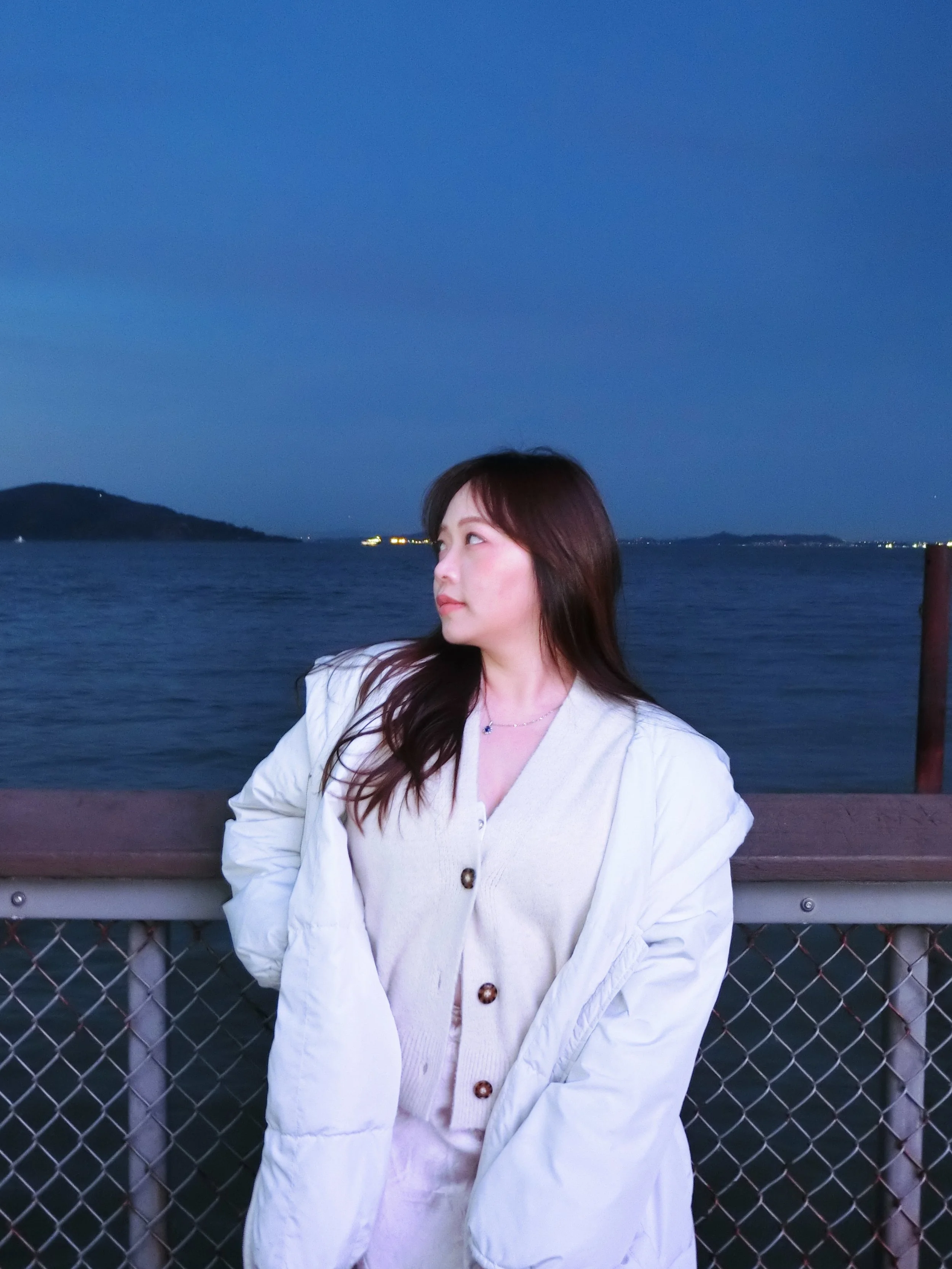 A woman standing on a waterfront pier at dusk, looking to her left with a calm ocean and distant landforms in the background.