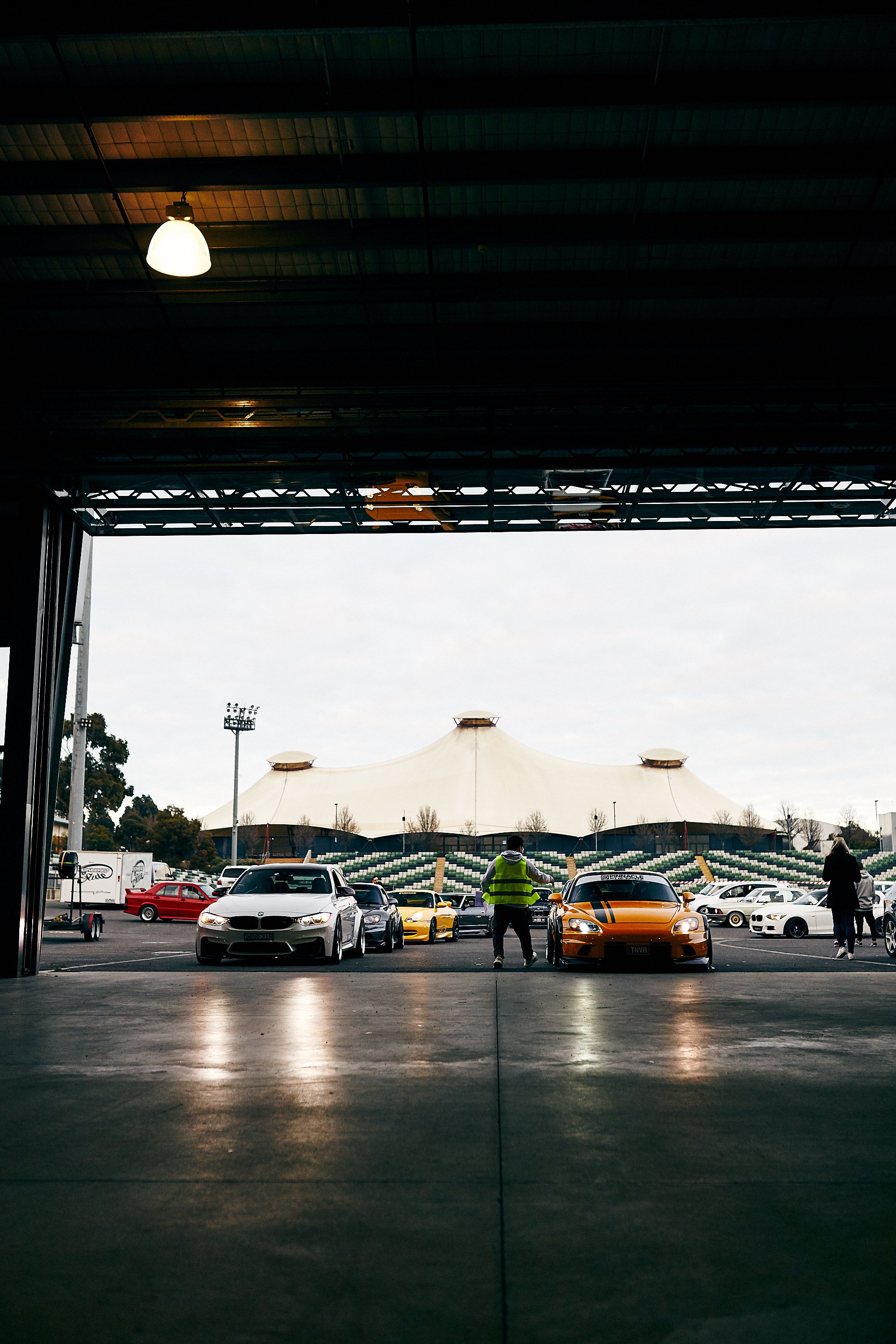 View of a car park with several sports cars, including a white one, a yellow one, and an orange one, parked in front of a large, tent-like stadium structure. A person in a high-visibility vest stands between the cars, and another person is taking a photo on the right. The photo is taken from inside a garage or covered area, looking outward.