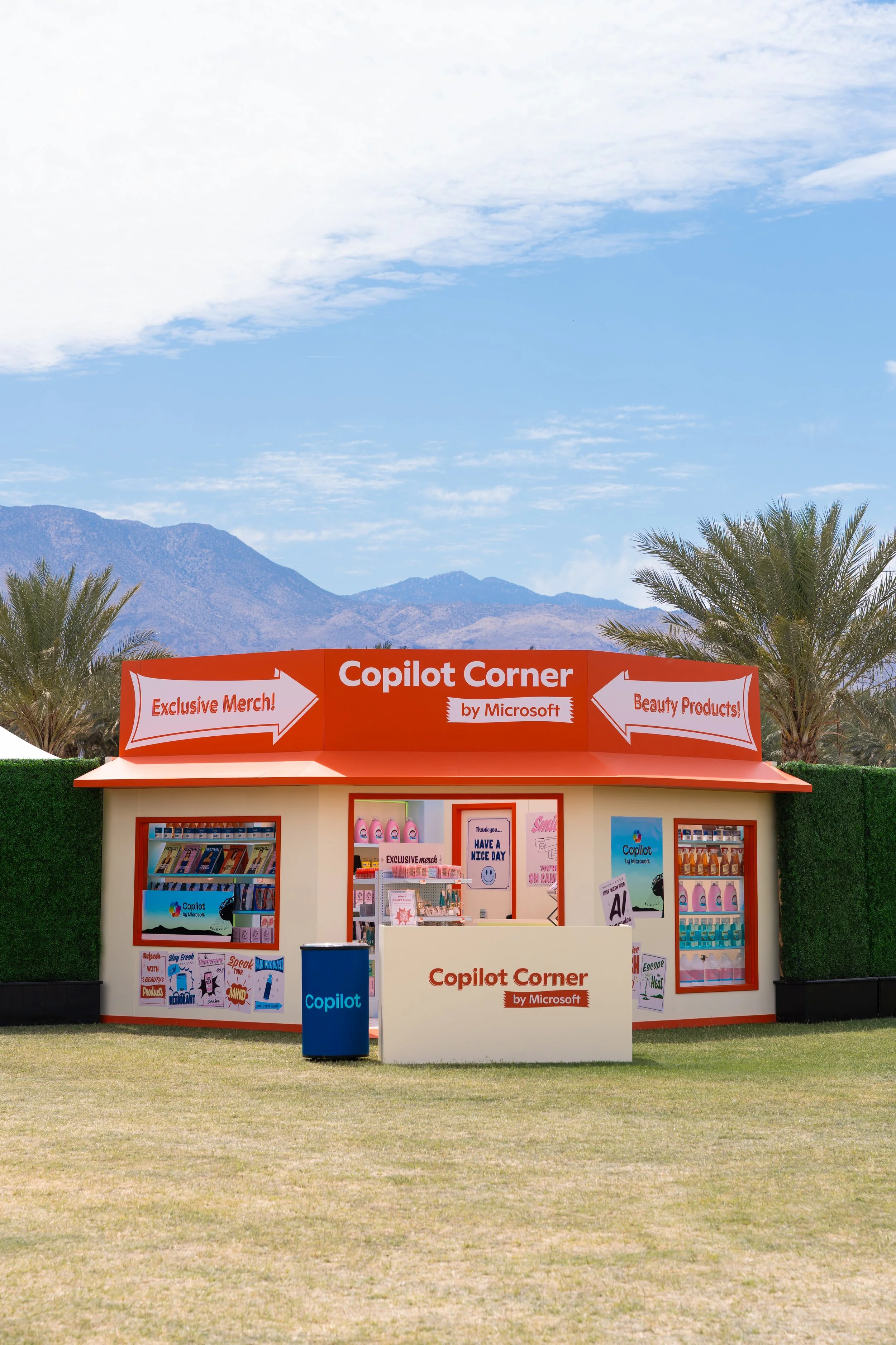 A small pop-up shop labeled 'Copilot Corner by Microsoft,' offering merchandise, beauty products, and promotional material, set against a mountainous landscape with palm trees and blue sky.