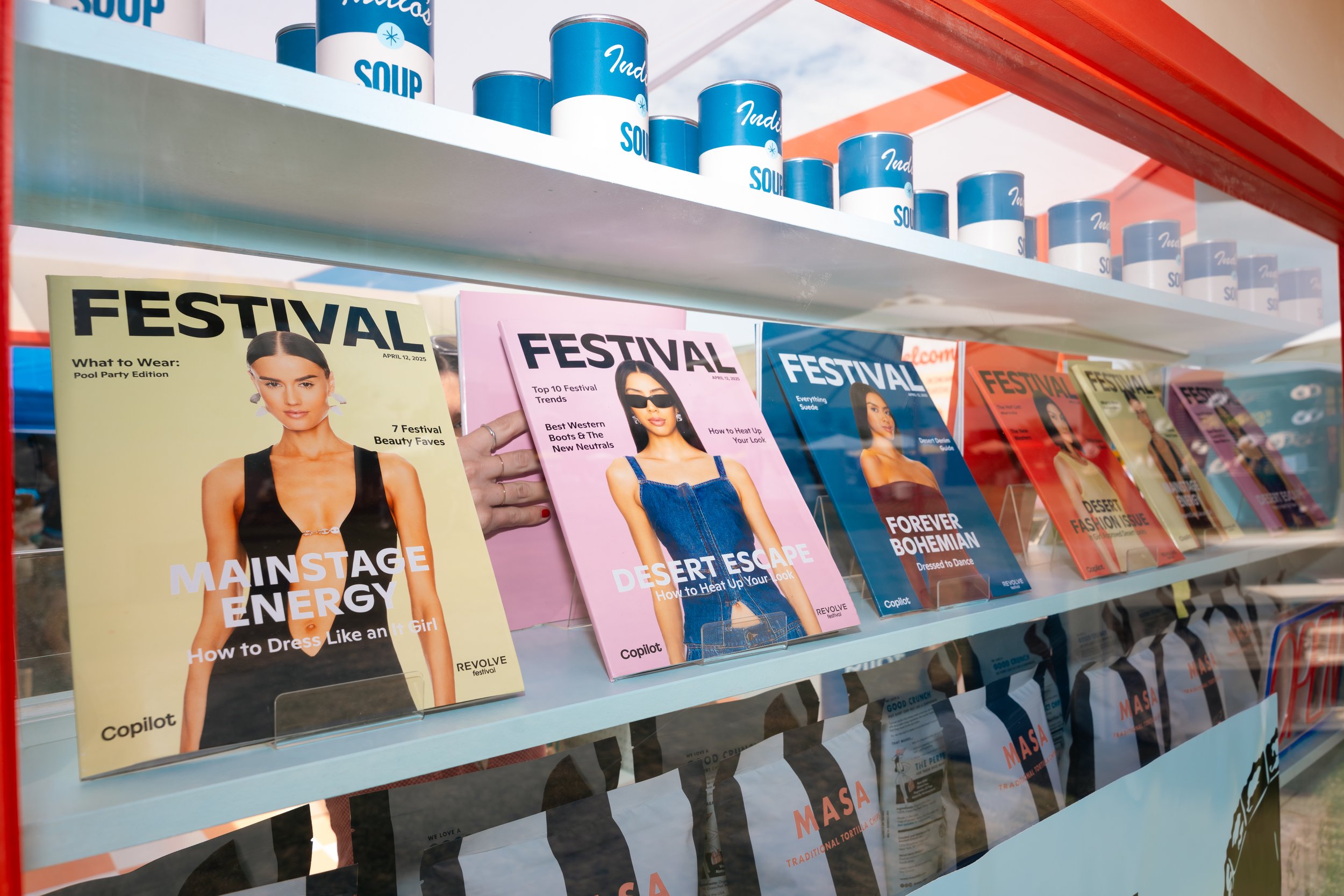 Magazines titled 'FESTIVAL' displayed on a shelf with various cover designs and colors, including yellow, pink, and blue. Cans of soup are arranged on the top shelf. A person is partially visible holding a magazine.