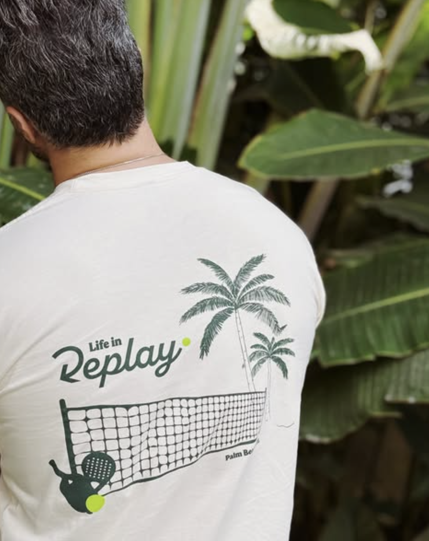 The back of a person wearing a white t-shirt with a graphic of palm trees, a tennis net, and the words "Life in Replay". The person is standing outdoors near large green tropical leaves.