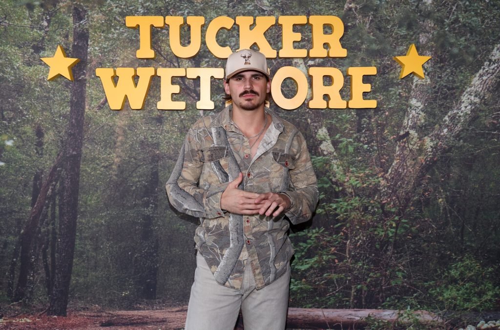 Person wearing camouflage clothing and a hat standing in front of a forest-themed backdrop with the words 'Tucker Wetmore' and star decorations.