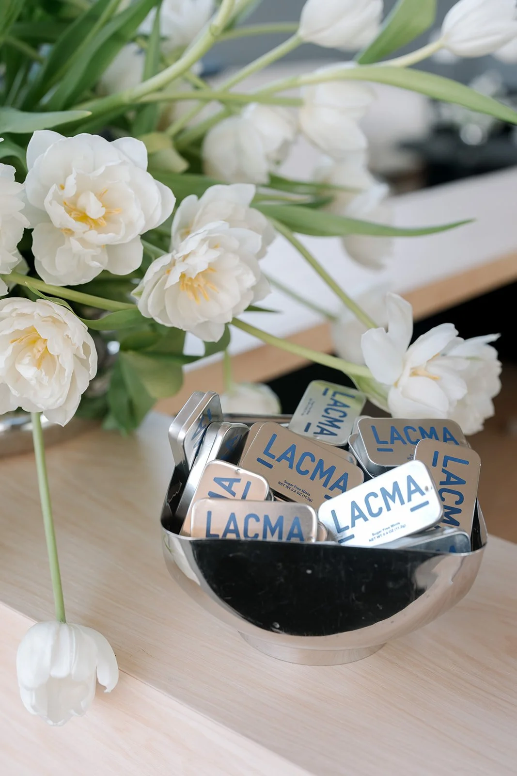 A bowl of LACMA-branded mints on a wooden surface with a bouquet of white tulips nearby.