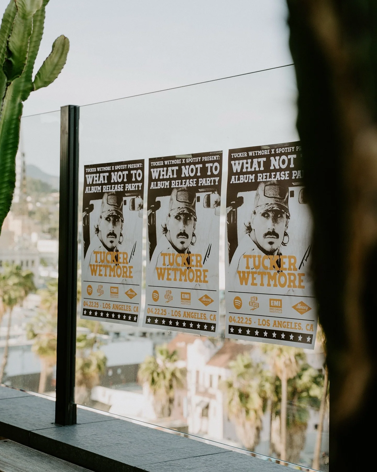 Three promotional posters for Tucker Wetmore's album release party, featuring a black-and-white image of a man with a mustache and a cap, with event details at the bottom. The posters are attached to a glass window with a city view in the background.
