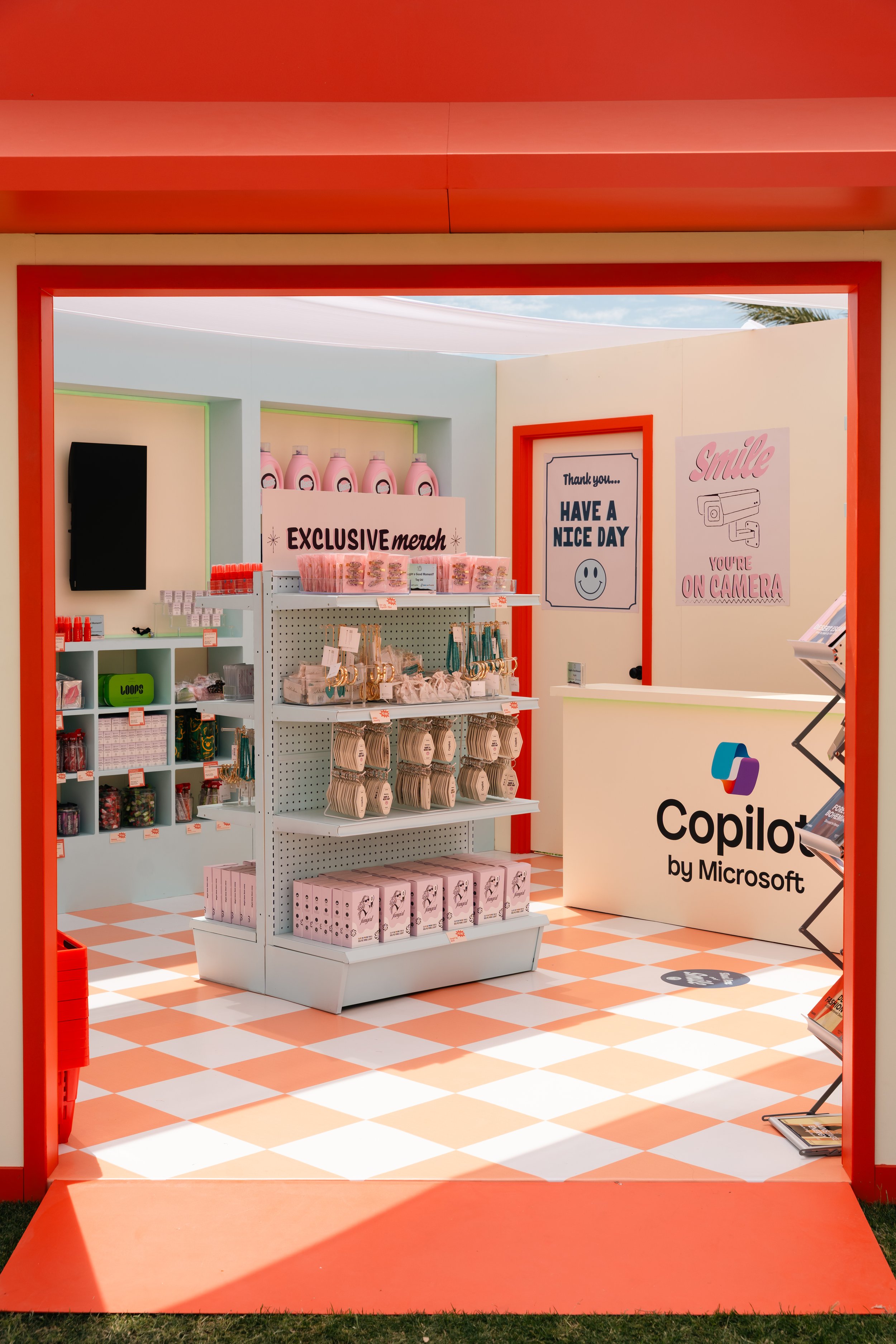 A brightly colored merchandise store with "Exclusive merch" sign and various products on shelves. The floor is checkered orange and white. Signs on the walls include "Thank you... Have a nice day" and "Smile, you're on camera." A "Copilot by Microsof
