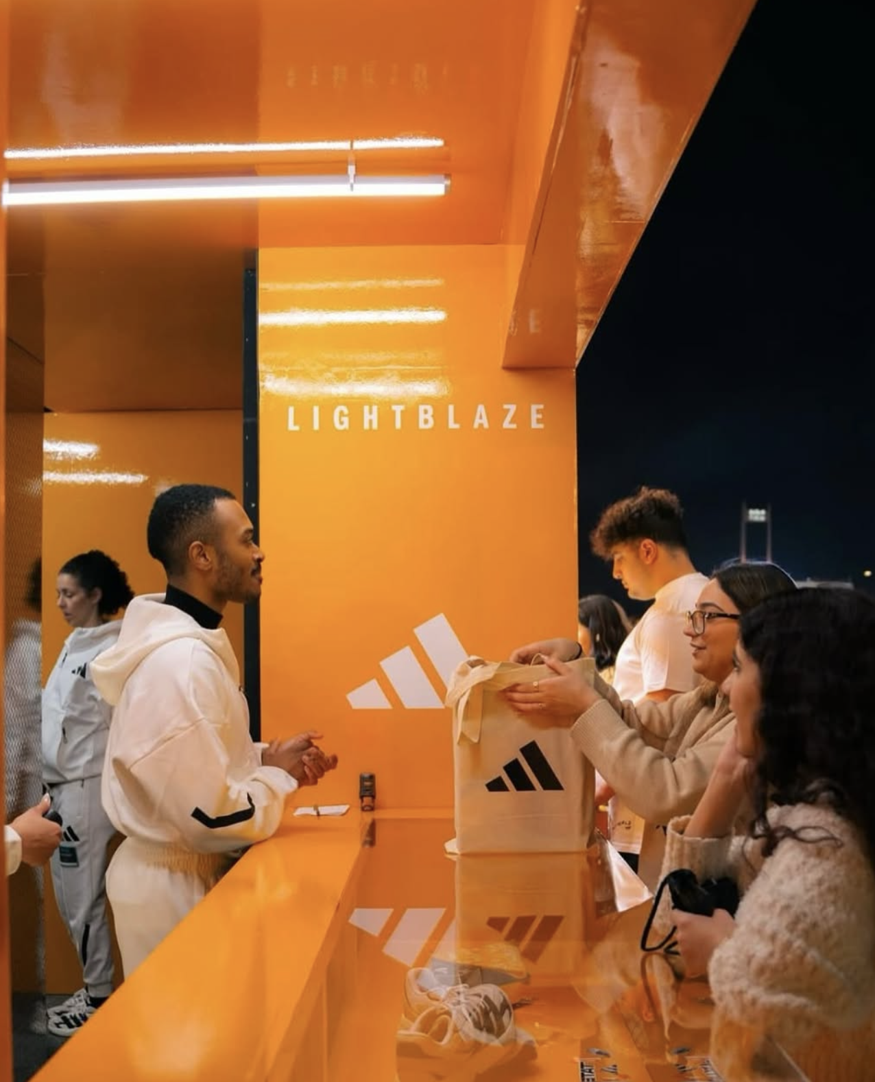 People at an Adidas booth labeled 'LIGHTBLAZE,' with an employee handing a branded bag to a customer.