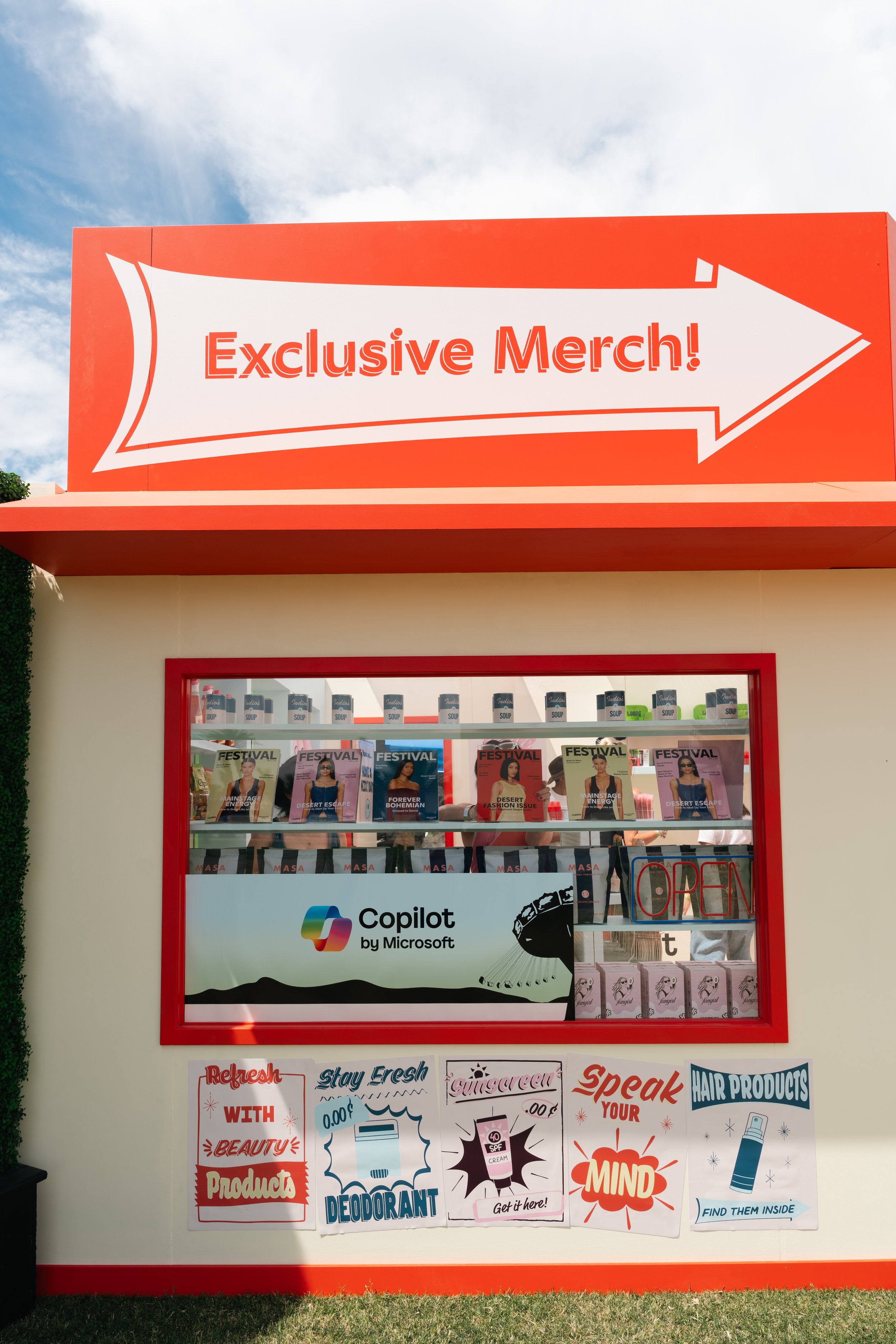 Booth displaying 'Exclusive Merch' sign with products visible in the window, including magazines and hygiene items. Promotion for Microsoft's Copilot and various product ads below the window. Clear sky background.