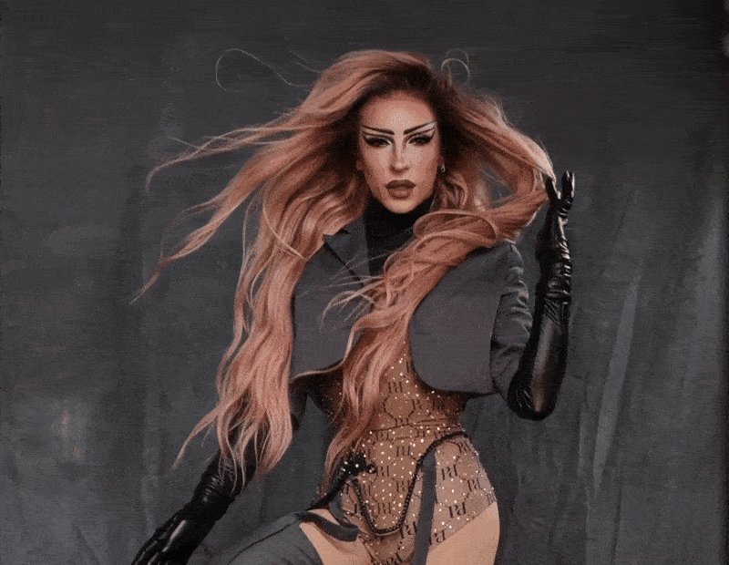 A woman with long, windblown reddish hair, wearing black gloves, a dark jacket, and revealing lingerie with a pattern, posing against a dark backdrop.