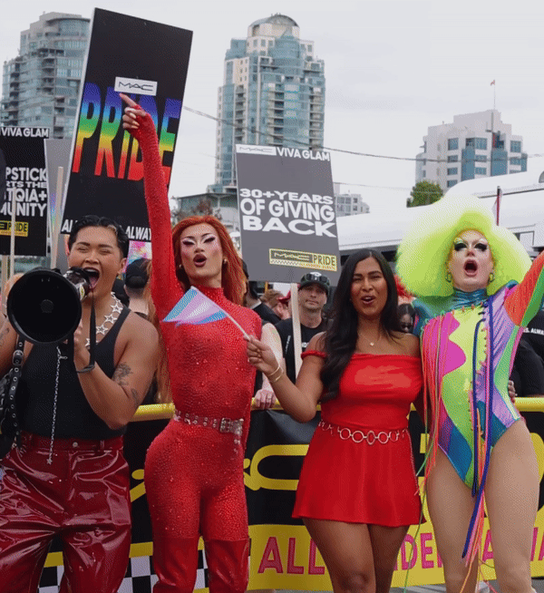 Four people celebrating at a Pride parade, holding a rainbow flag, with signs and city buildings in the background.