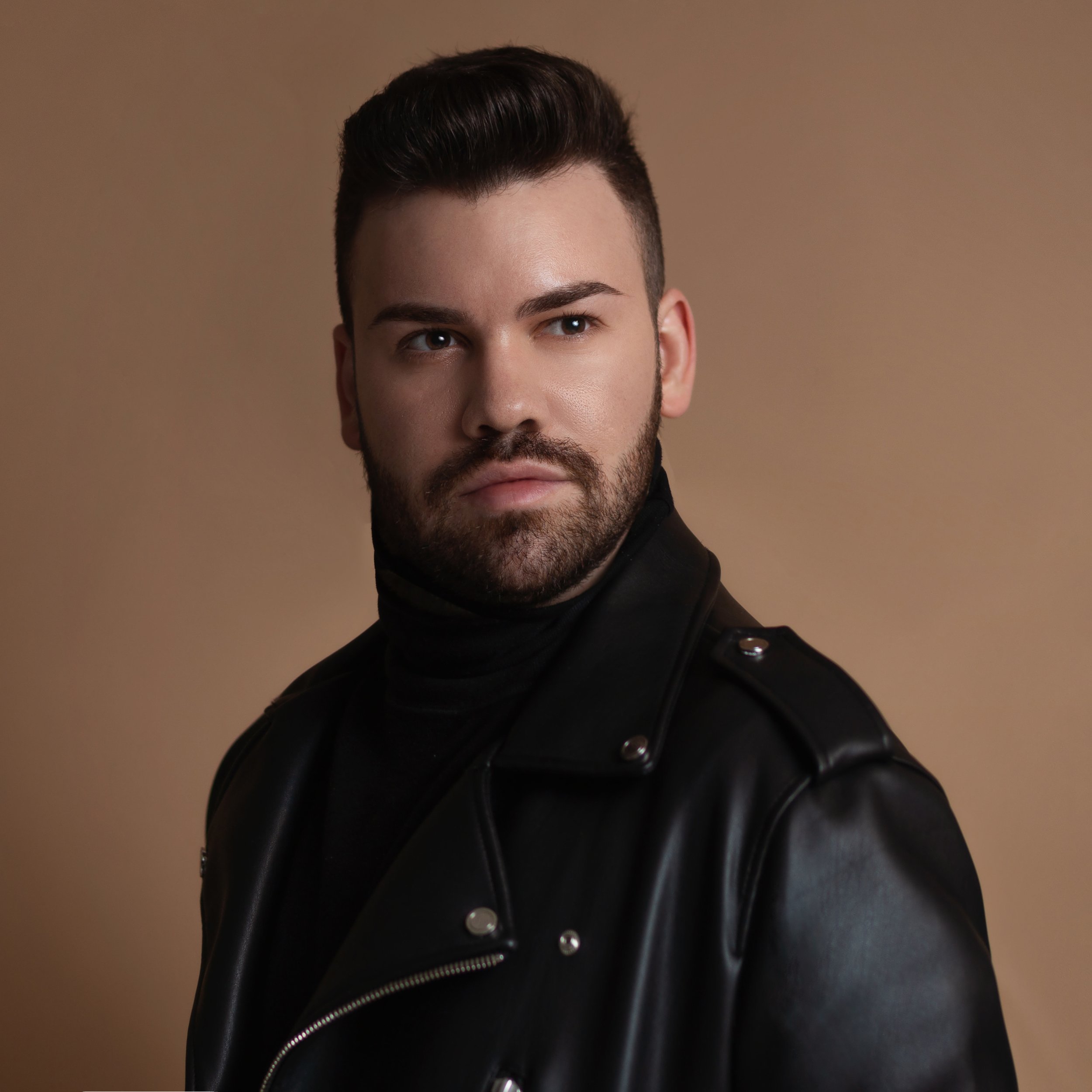 The Drag Series, Fernando Cysneiros, Portrait of a man with dark hair and a beard, wearing a black leather jacket and turtleneck, against a plain background.