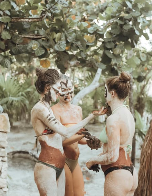 Three women with mayan clay on their bodies and faces, wearing swimsuits, standing outdoors under a tree, engaging in a clay ceremony or playful activity.