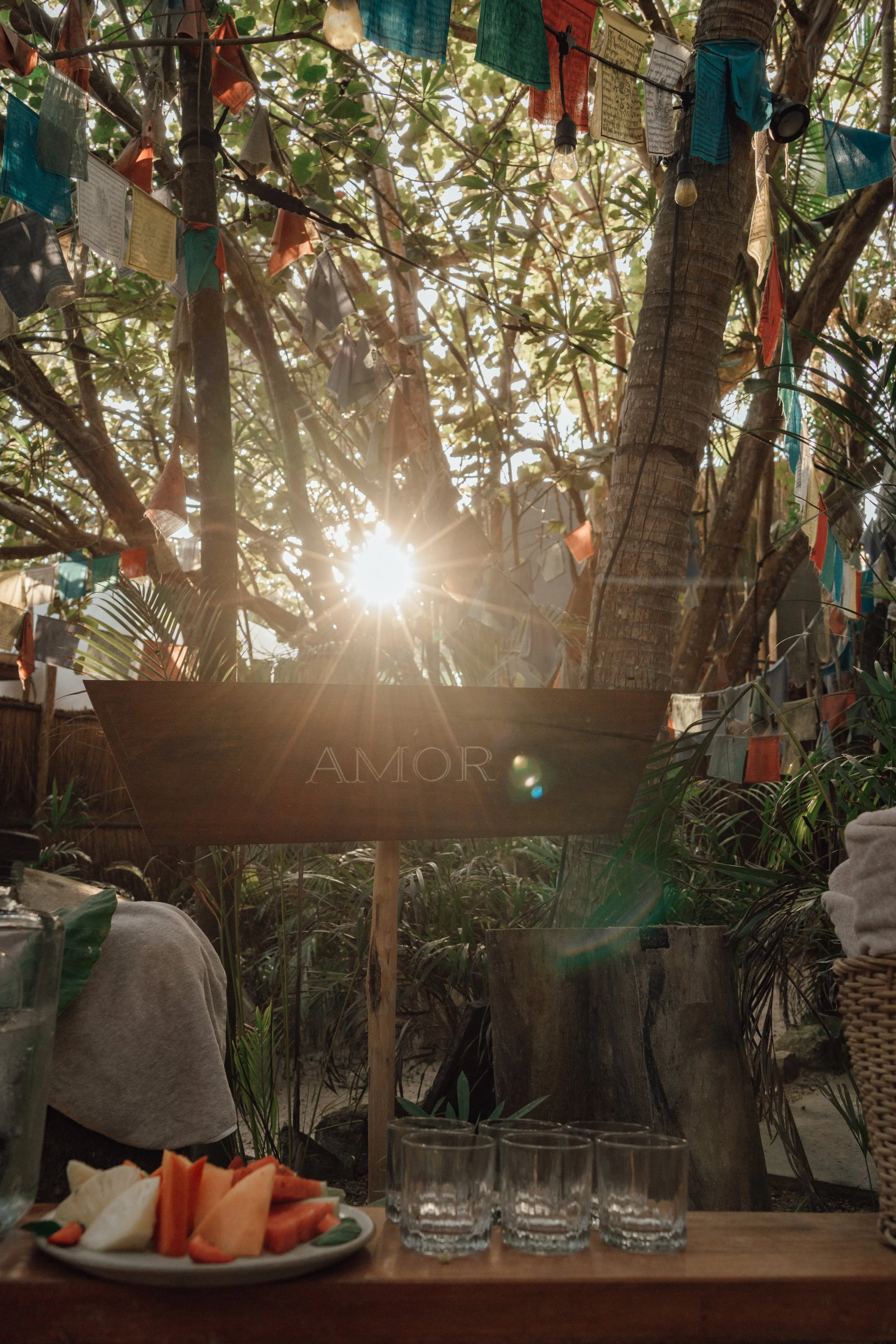 Sunlight filters through trees with colorful prayer flags, a wooden sign reads 'Amor', a plate of fruit, and glasses on a rustic table, suggesting an outdoor setting or celebration.