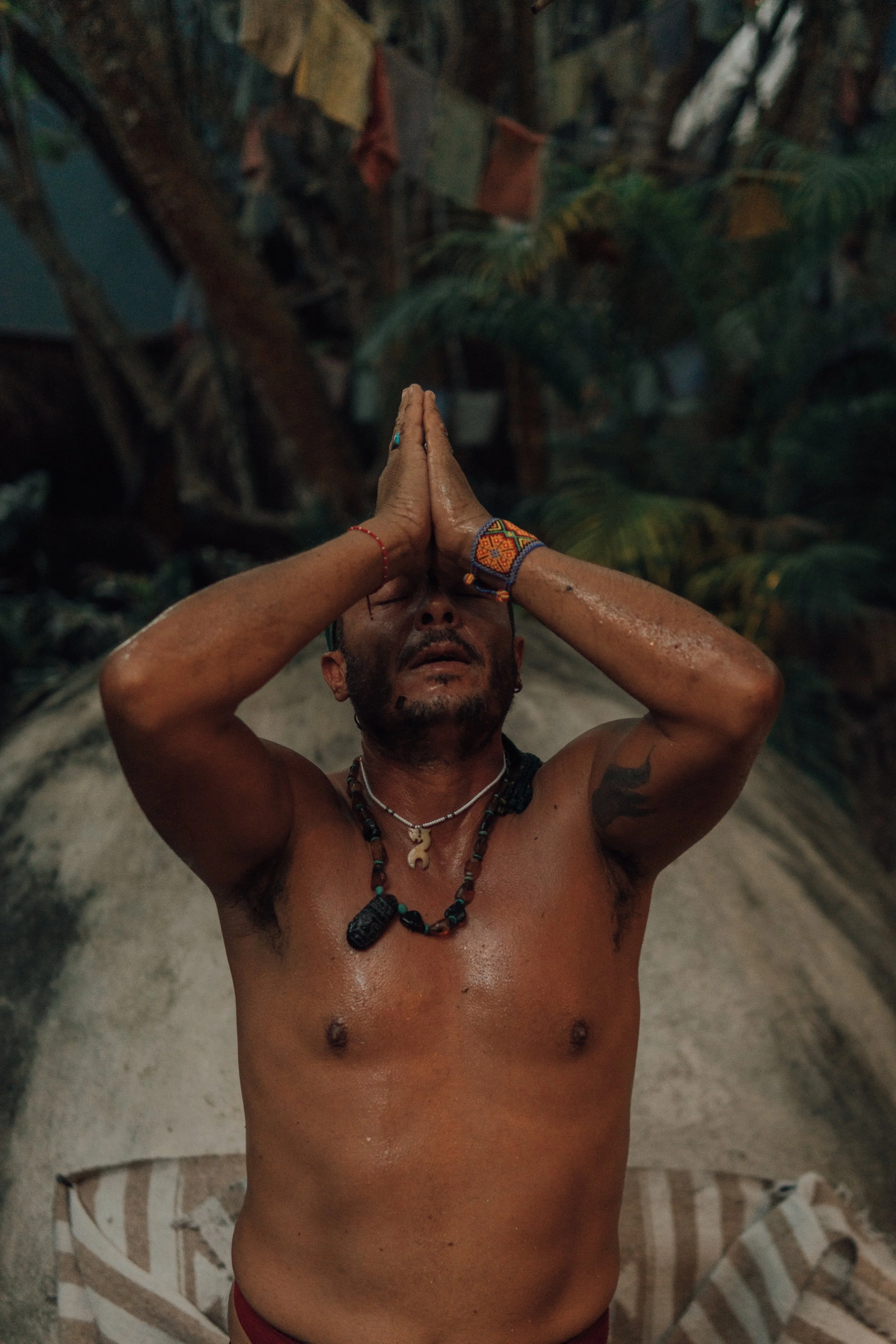 A shirtless man with tattoos and jewelry, in a spiritual or meditative pose with hands pressed together above his face, outdoors after the ending of a Medicine Wheel Temazcal