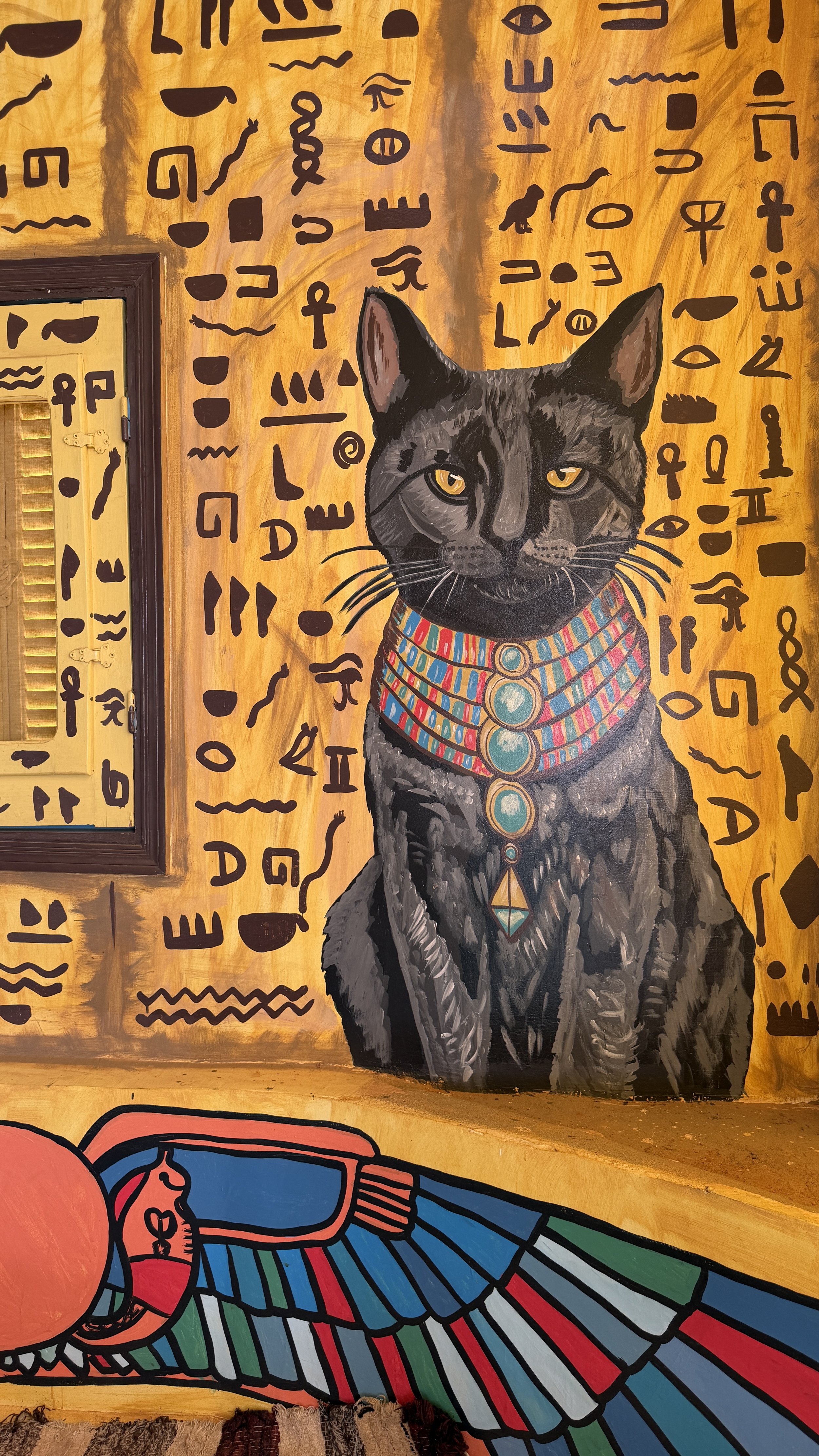 A mural of a black cat wearing colorful Egyptian-style jewelry, with Egyptian hieroglyphics painted in black on a yellow-orange background behind it.