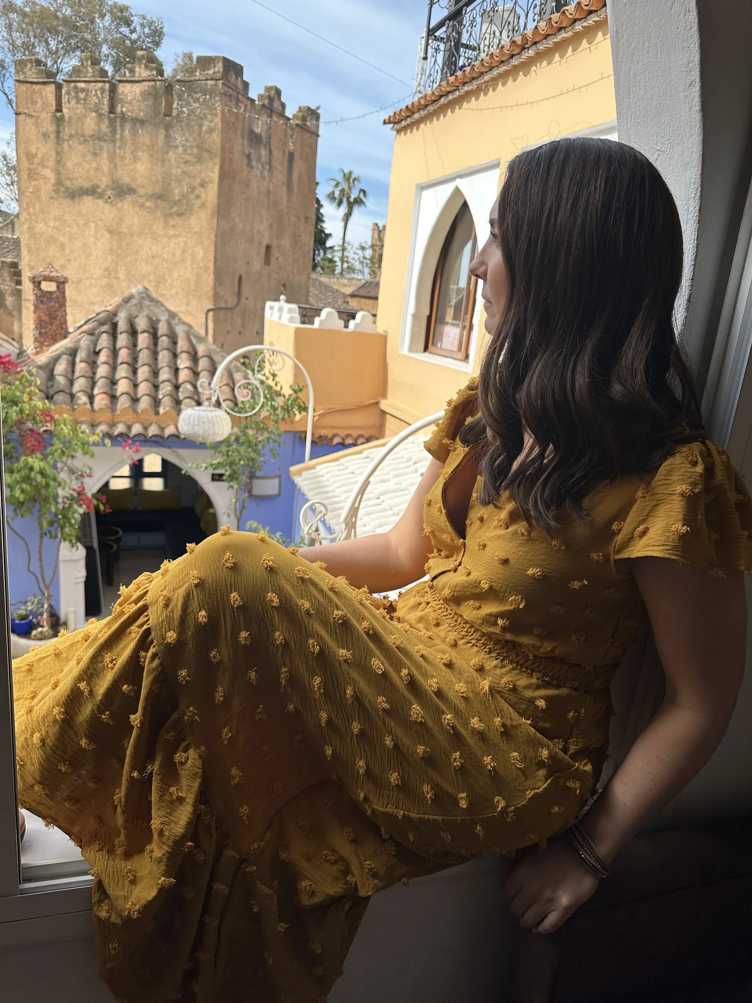 Woman in flowing yellow dress with textured pattern sitting on balcony overlooking colorful Moroccan architecture with terracotta roofs and ancient castle walls in Chefchaouen