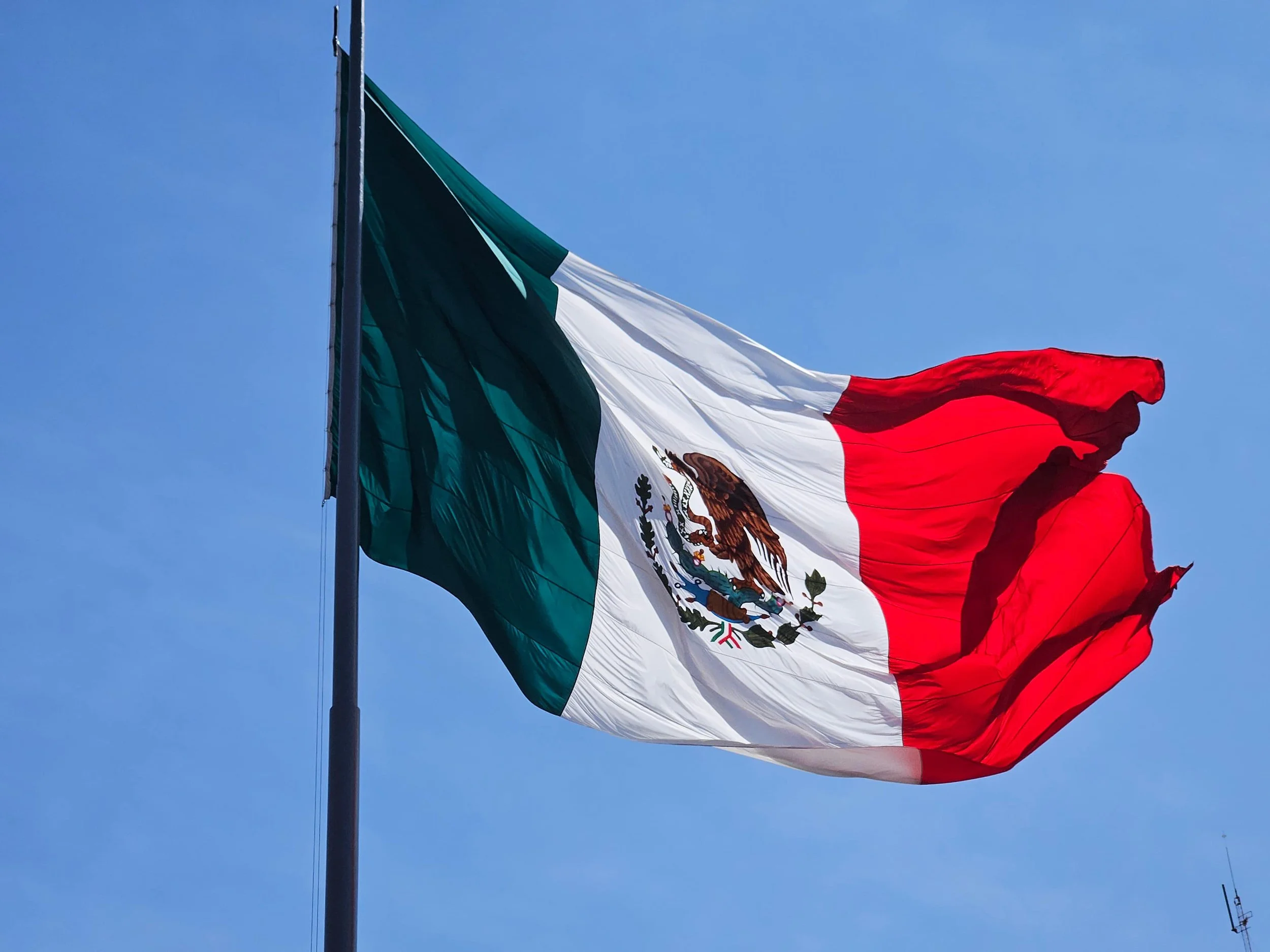 Mexican flag waving against a clear blue sky, displaying the green, white, and red vertical stripes with the national coat of arms featuring an eagle in the center.