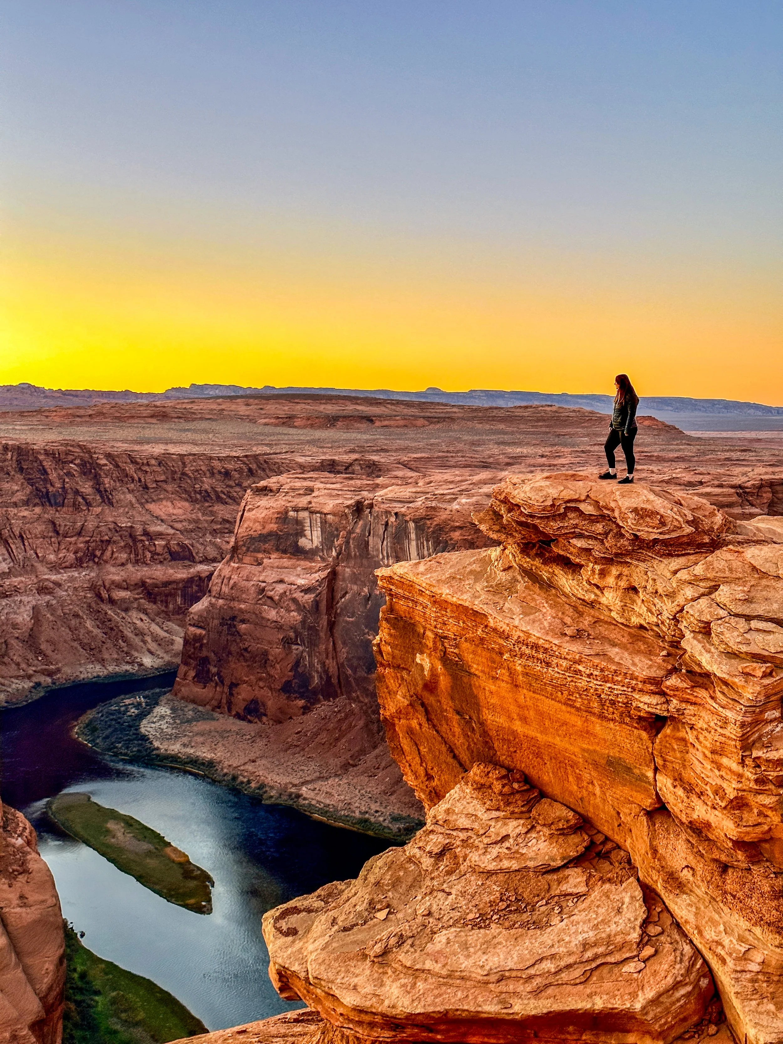 A person standing on the edge of a rocky cliff overlooking the Colorado River in a canyon at sunset.