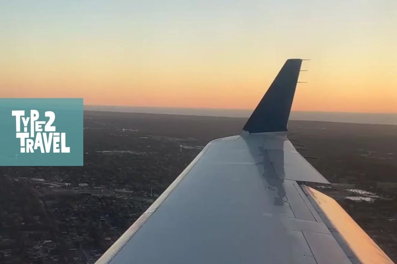 View from airplane window showing wing silhouetted against golden sunset sky over landscape below, capturing the beauty and uncertainty of travel, Type 2 Travel podcast logo in corner