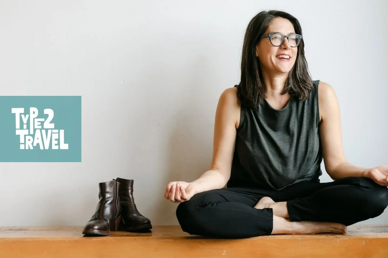 Woman in black sitting cross-legged in meditation pose on wooden platform with Type 2 Travel podcast logo