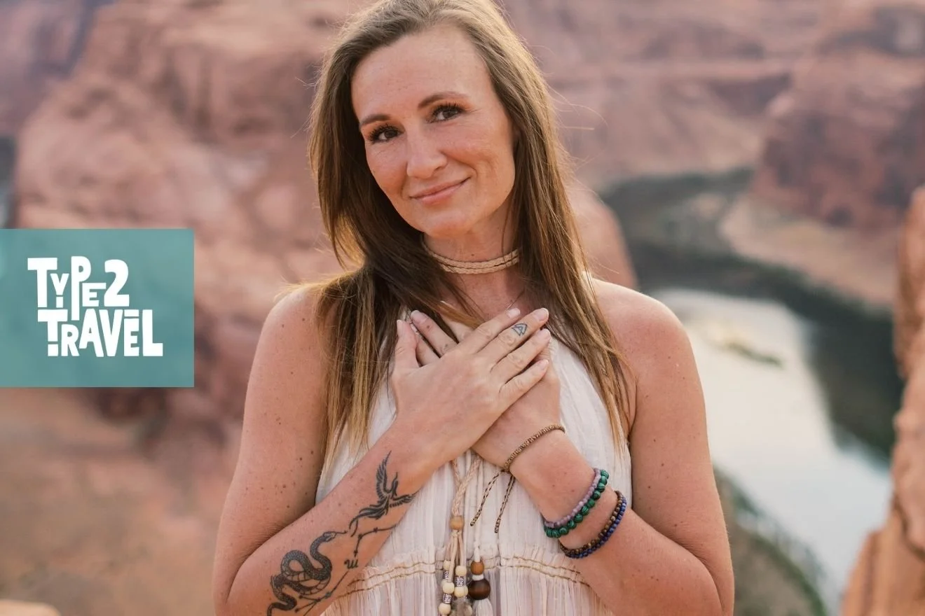Woman with long brown hair, hand over her heart, wearing a white halter top with beaded bracelets and a visible tattoo on her arm, photographed outdoors with a desert/canyon landscape in the background.