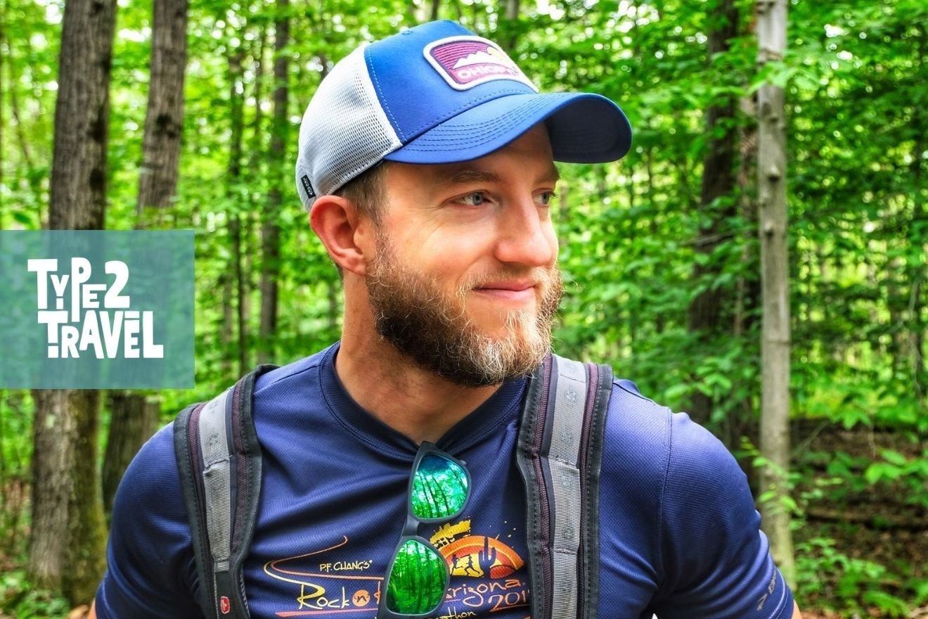 Joe Baur wearing a blue trucker hat and backpack, smiling in a lush green forest setting, with Type 2 Travel podcast logo in corner