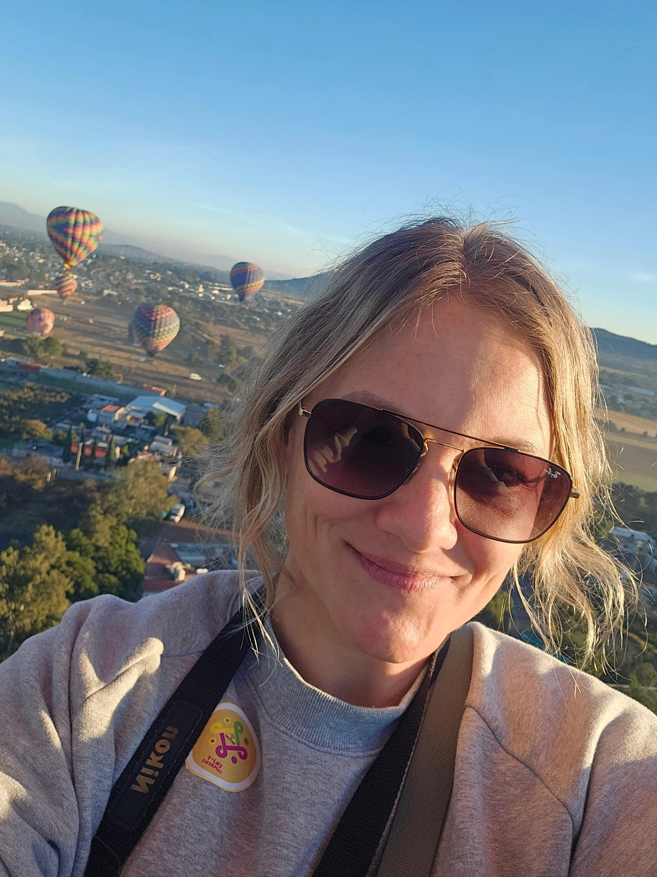 Woman wearing sunglasses taking a selfie with hot air balloons floating in the distance over the landscape of Mexico during golden hour sunrise or sunset.