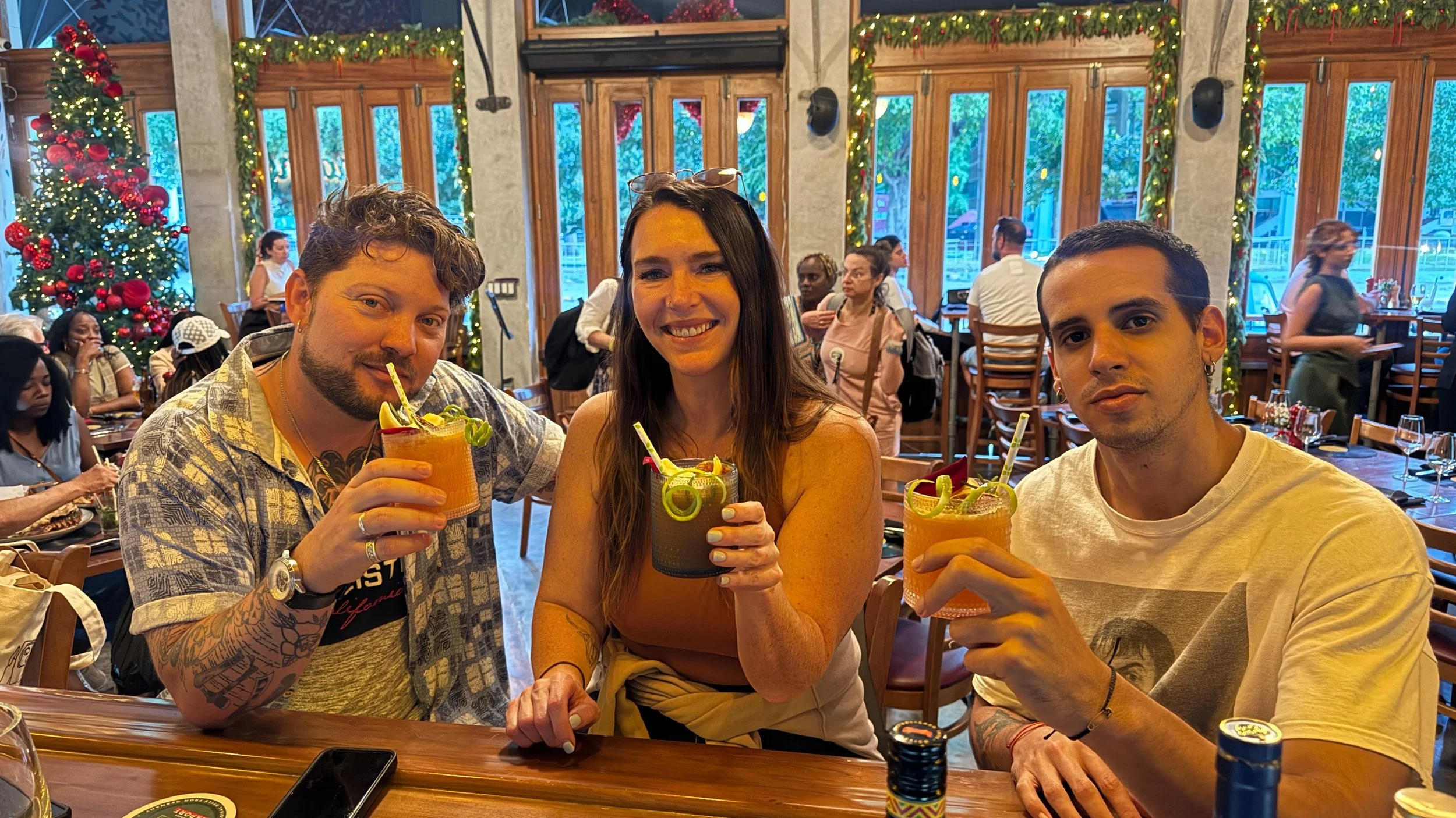 Three travelers enjoying cocktails at festive restaurant bar decorated with Christmas lights and tropical decor