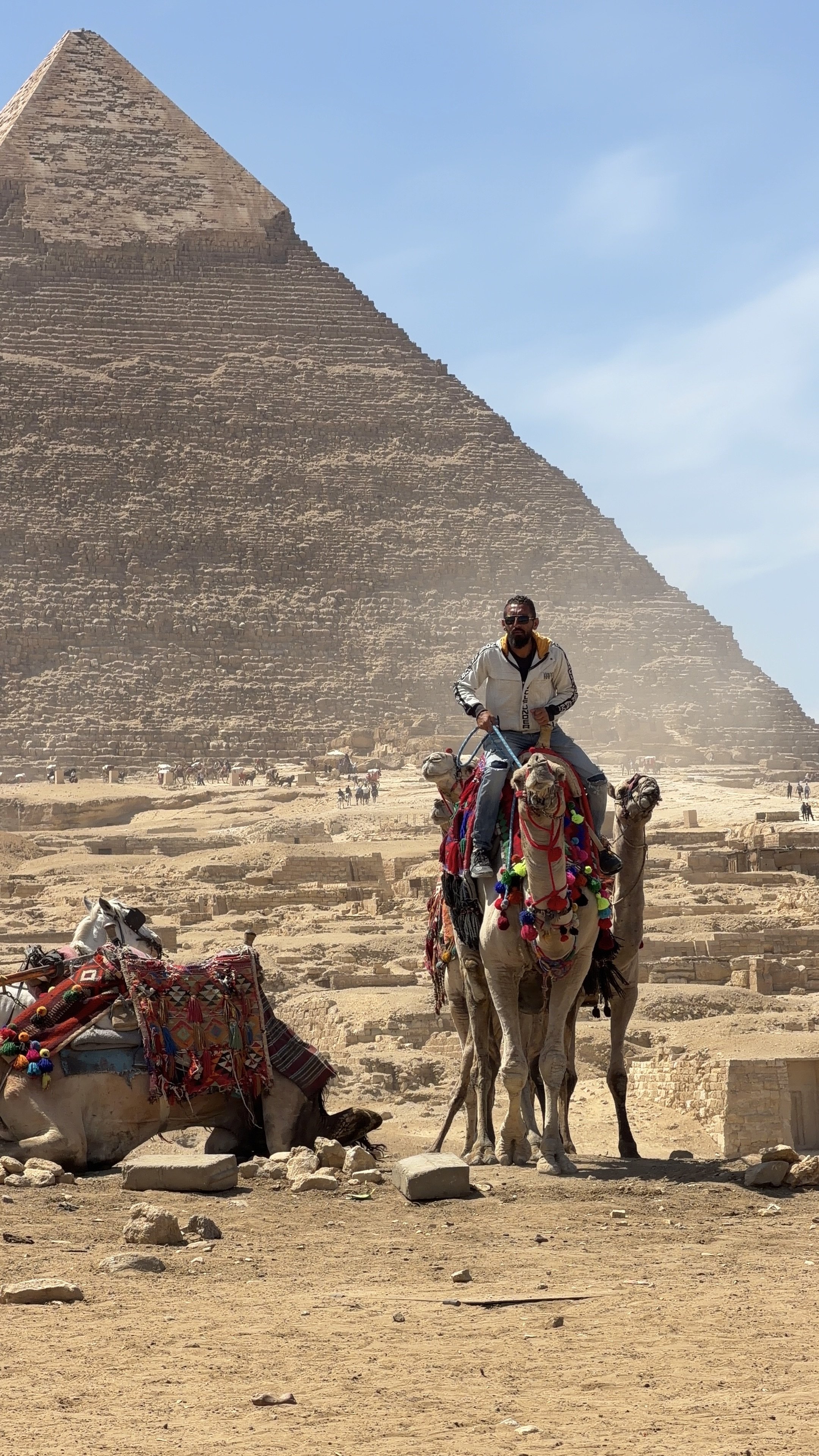 A man riding a camel in front of the Great Pyramid of Giza in Egypt, with other camels and tourists in the background.