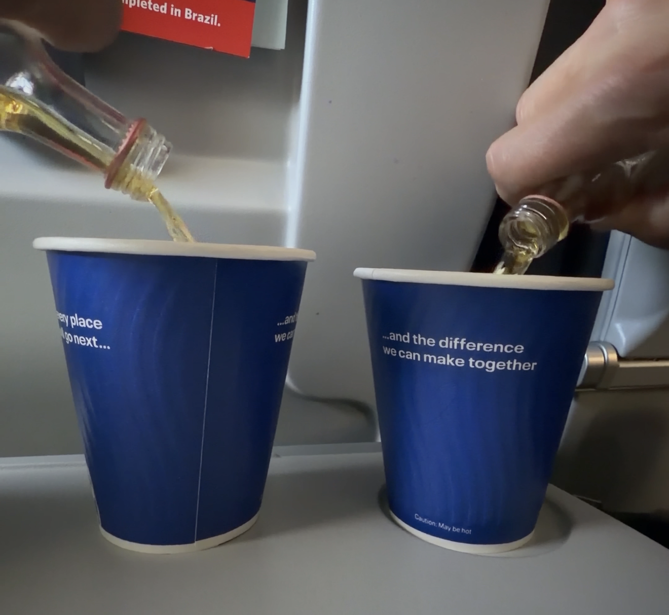 Two hands pouring mini bottles of liquor into blue paper cups on an airplane tray table.