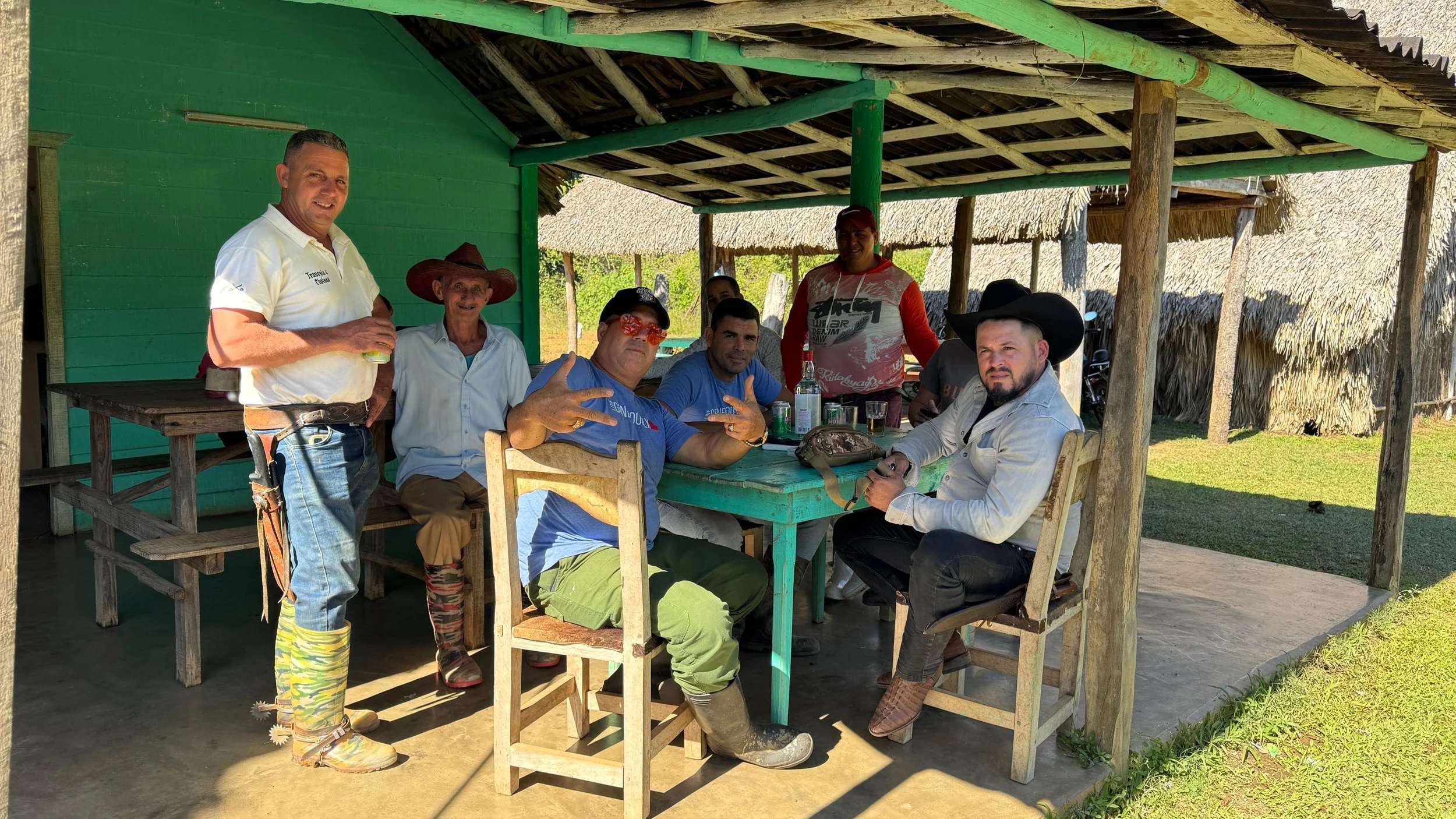 Group of Cuban locals and travelers gathering at rustic outdoor palapa with green wooden beams and thatched palm roof in rural Cuba, sharing drinks and conversation around simple wooden table