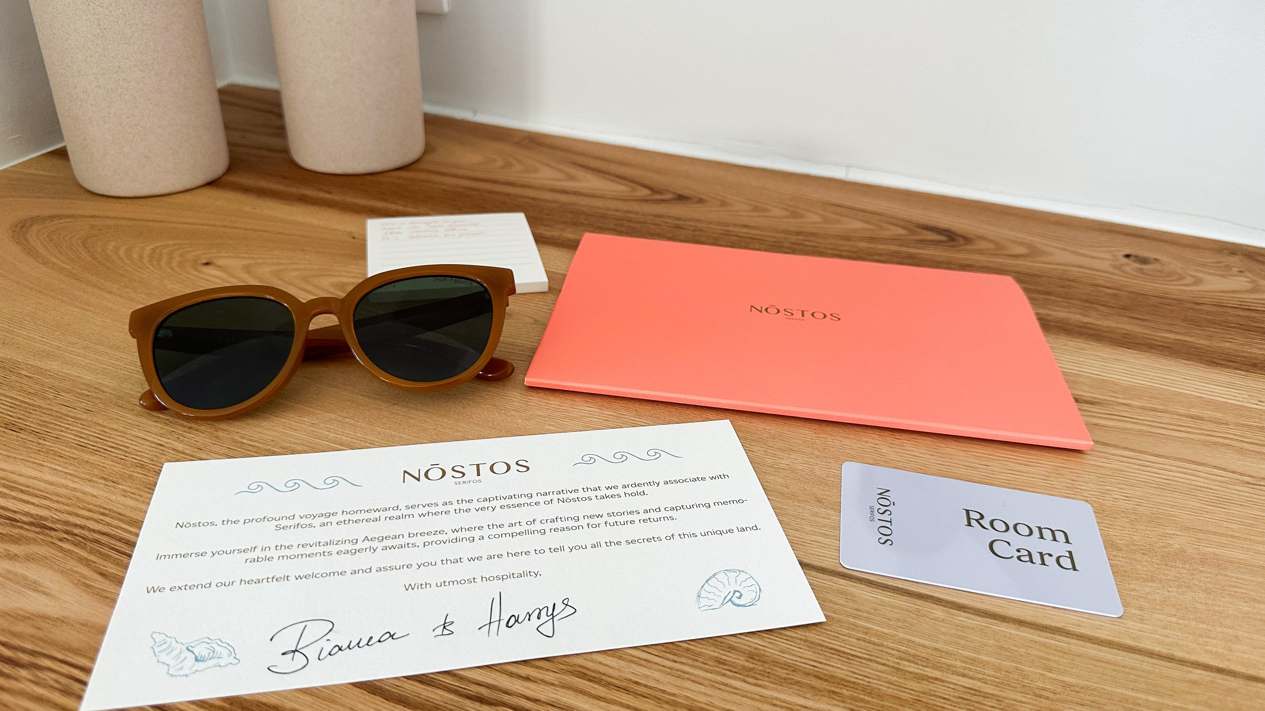 A wooden surface displaying Nostos branded items including a coral-colored folder, welcome letter, room key card, and stylish sunglasses, conveying a thoughtful and curated guest experience.